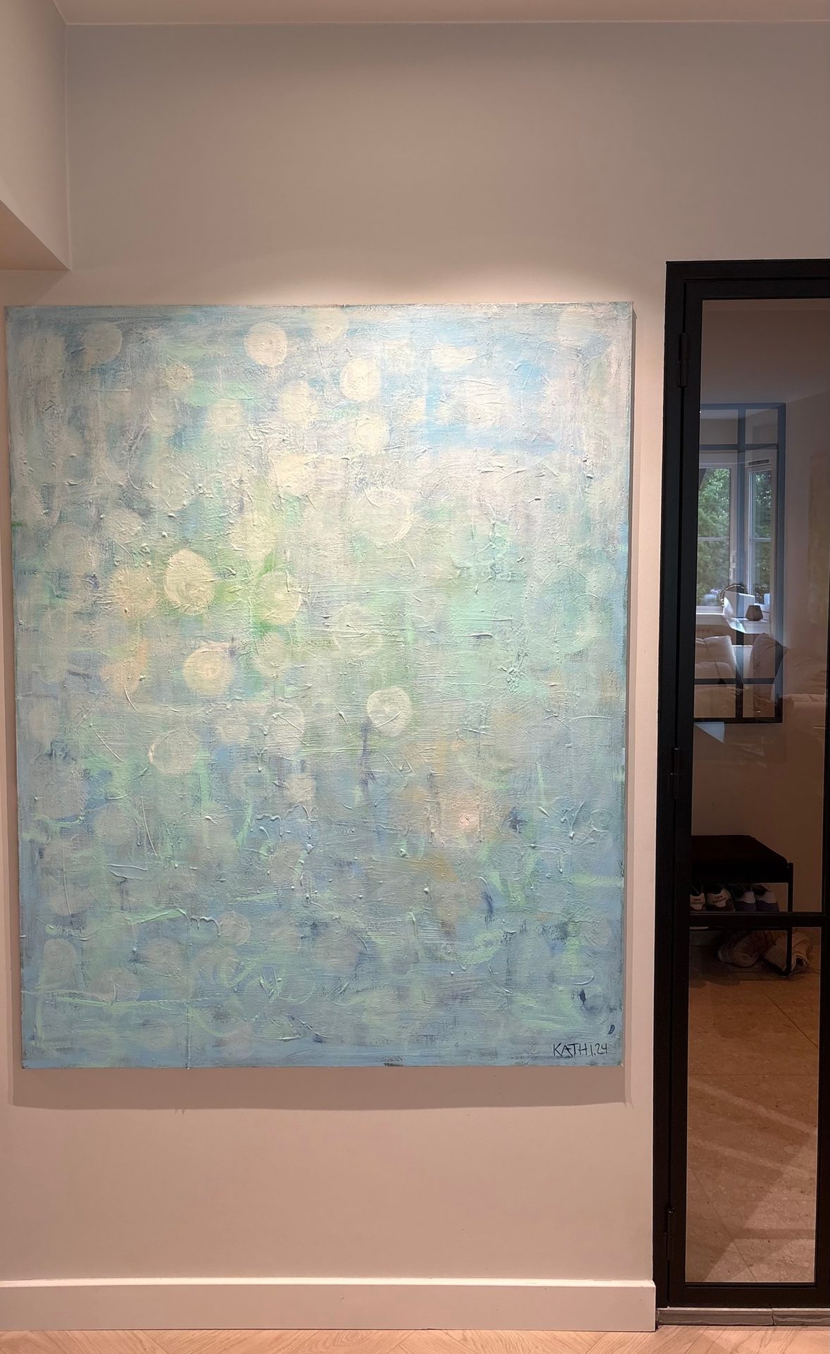 Abstract painting with light blue, green, and white circular shapes on a wall by a glass door.
