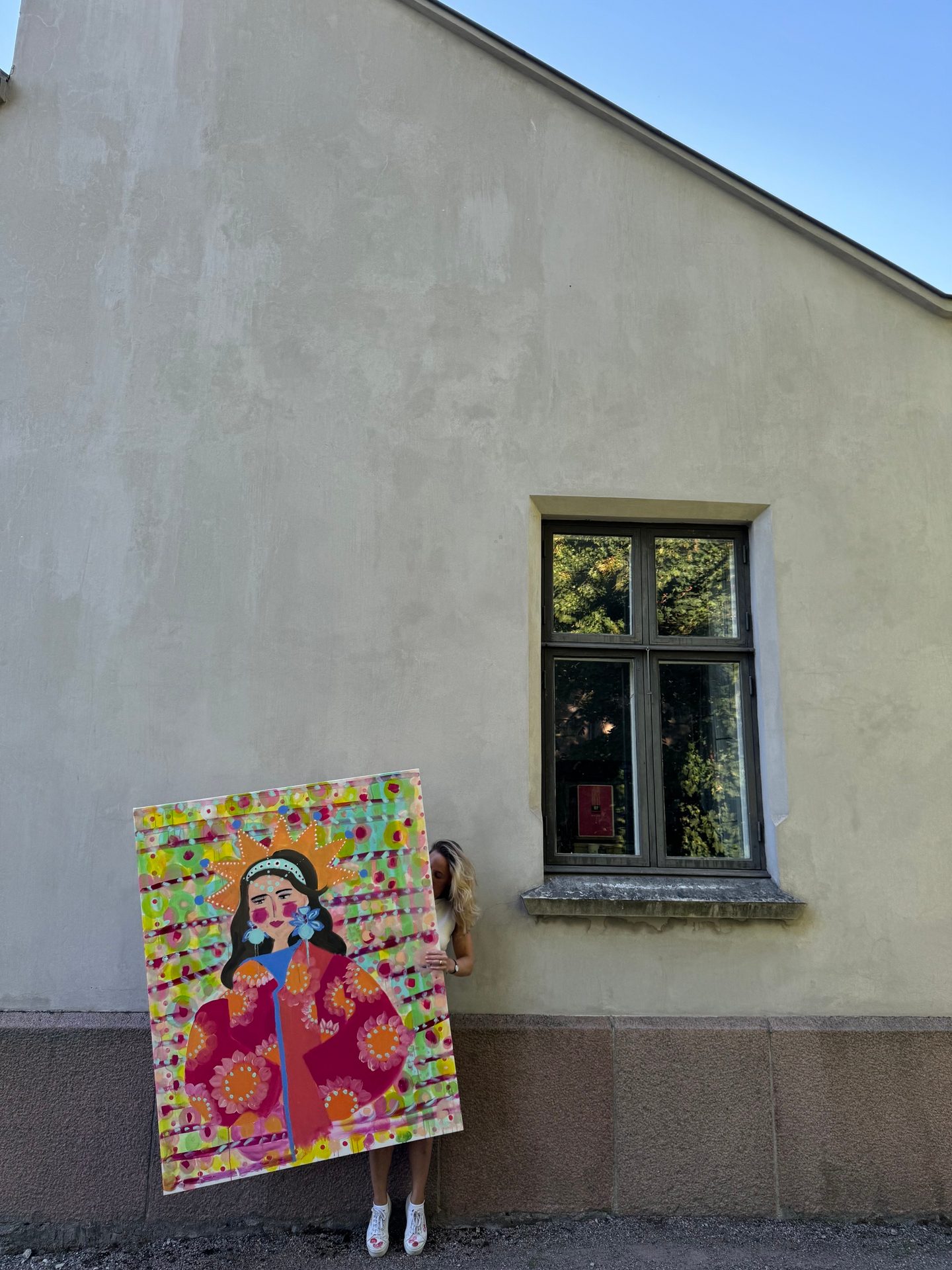 Woman holding a large, colorful painting of a queen-like figure by a building wall.