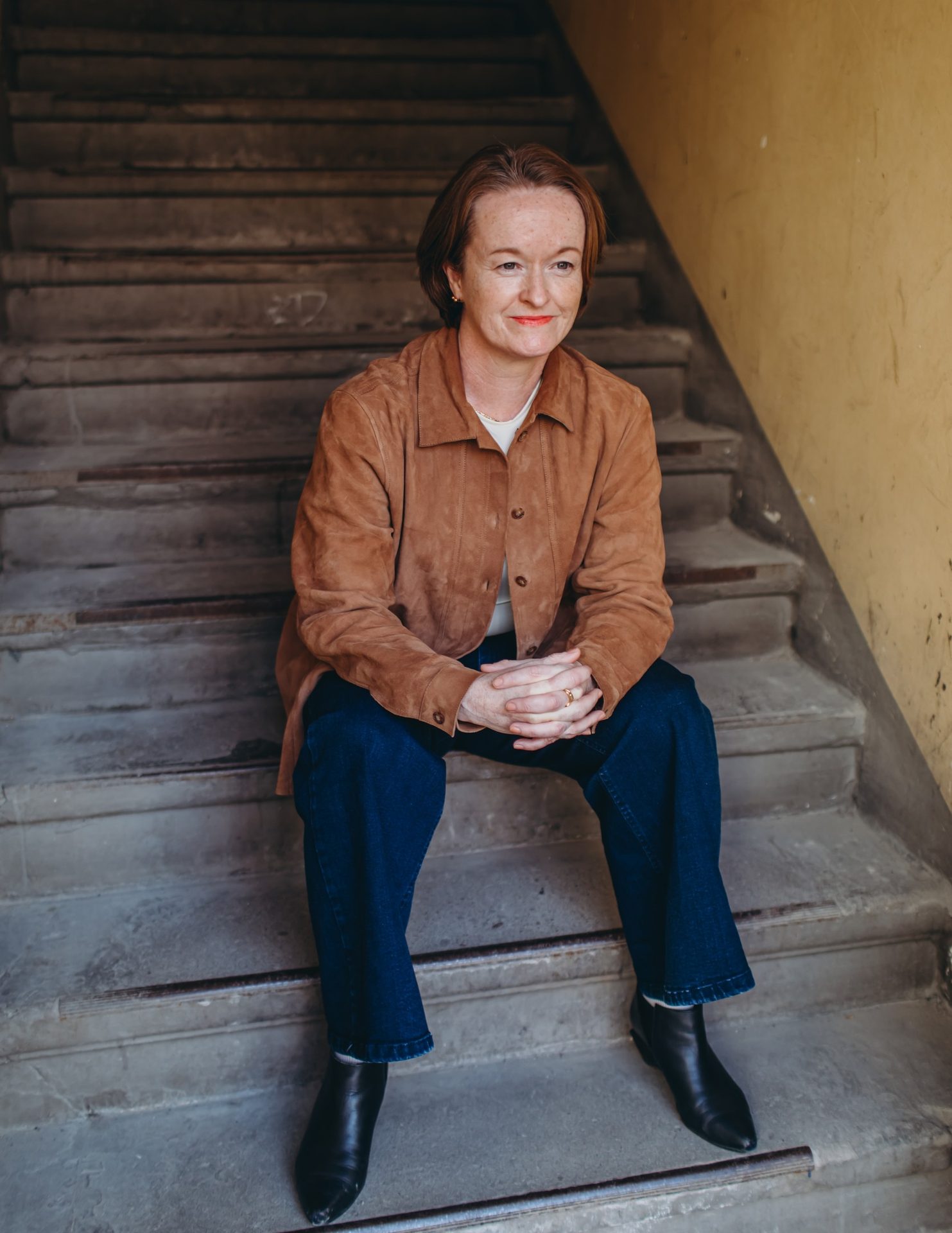 A woman with short brown hair and a brown jacket sits on concrete stairs, looking aside with a subtle smile.