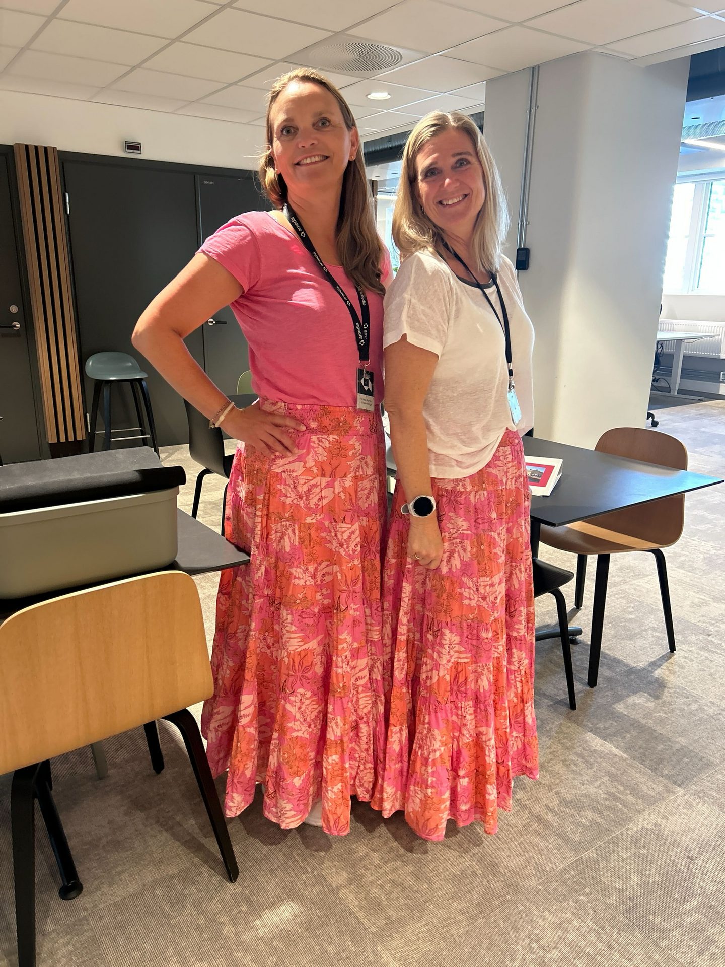 Two smiling women in matching floral skirts and t-shirts stand indoors.