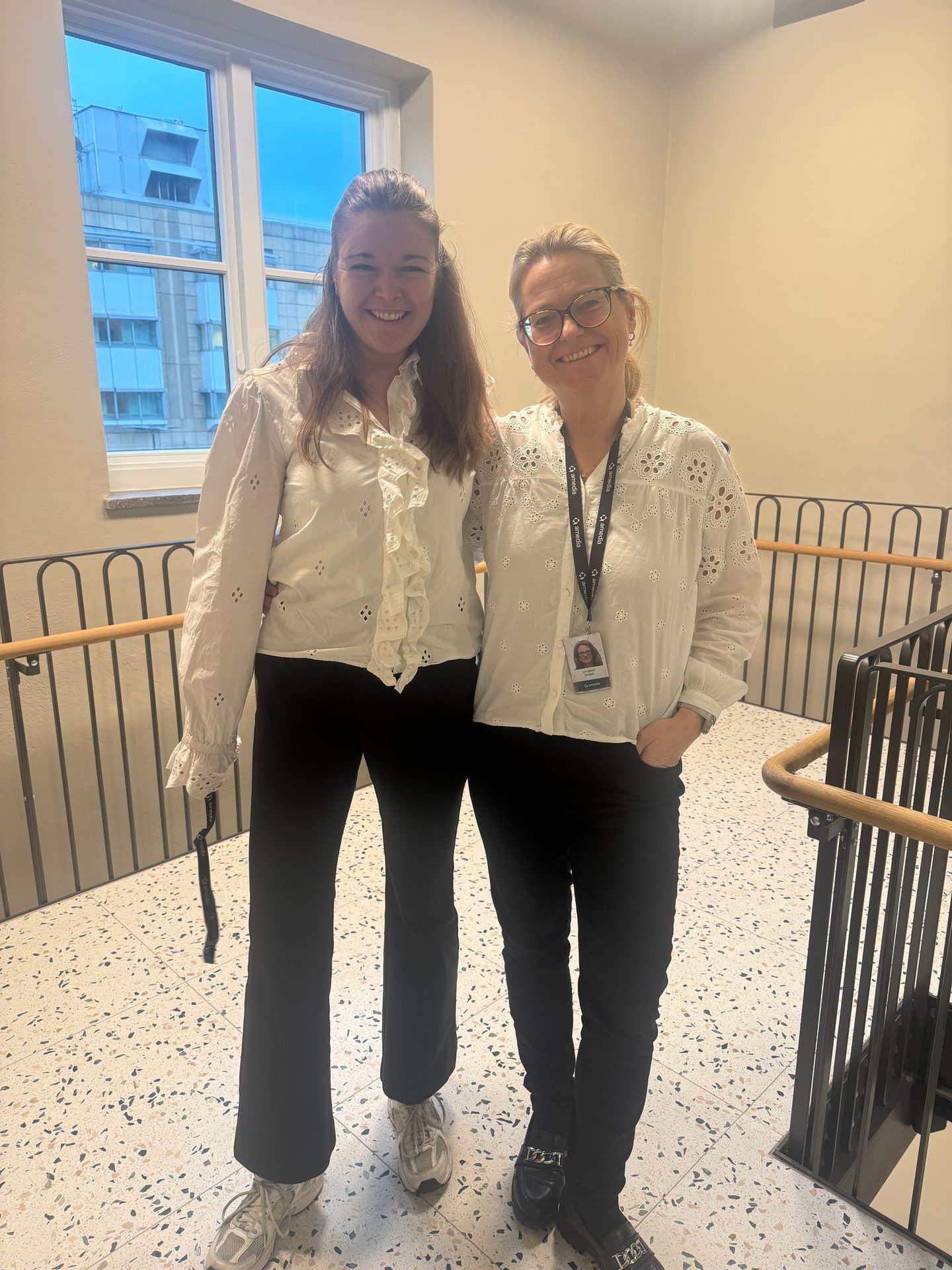 Two smiling women in white blouses and dark pants standing indoors near a window and staircase.