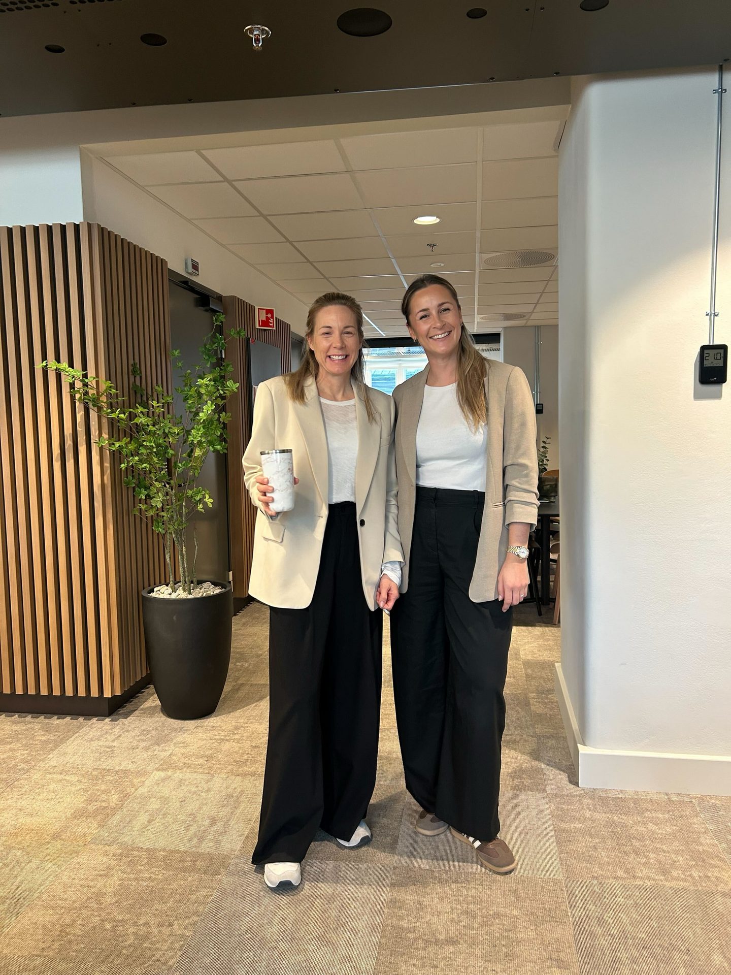 Two smiling women in blazers and black pants stand in an office hallway.