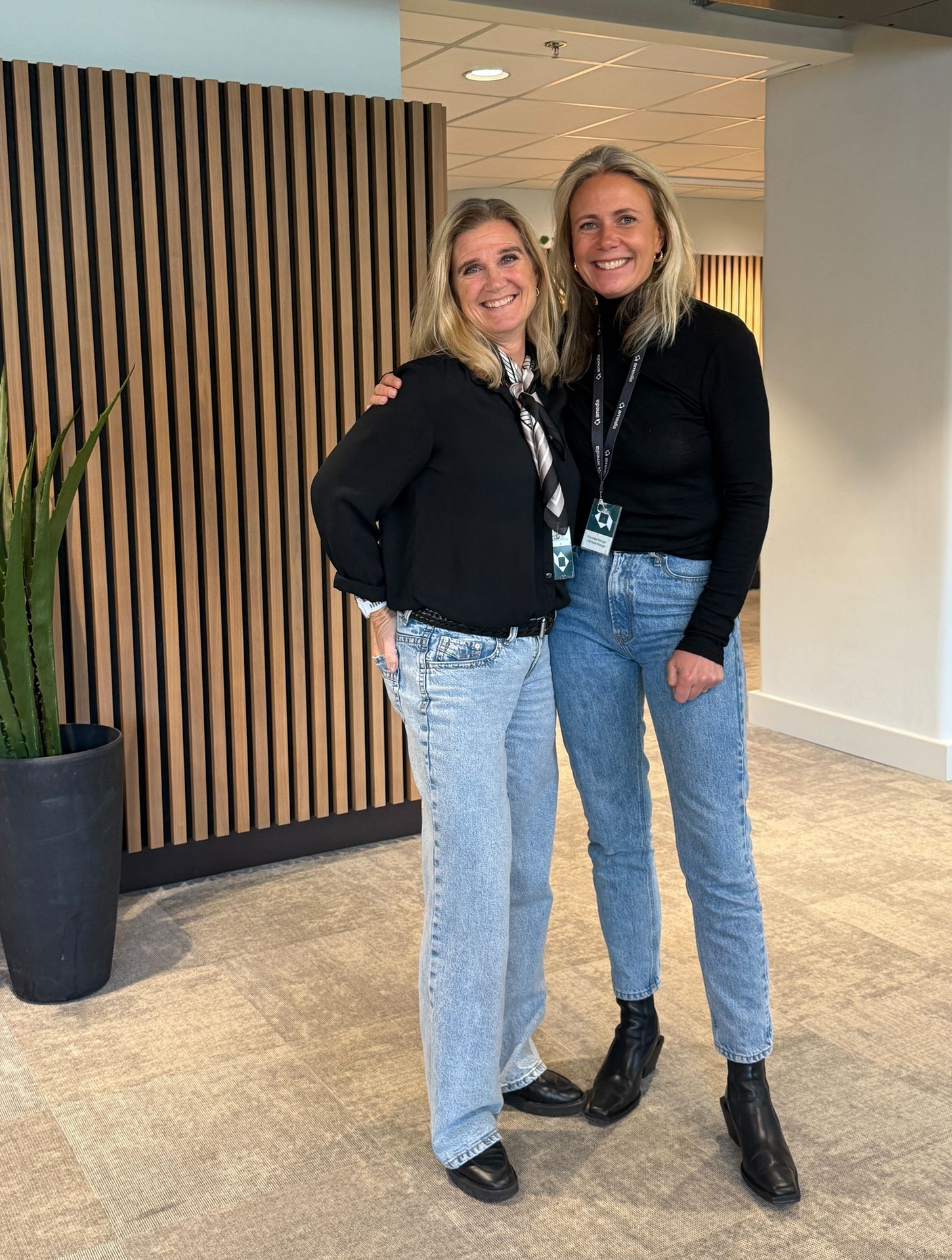 Two smiling blonde women in black tops and blue jeans pose in an office setting.