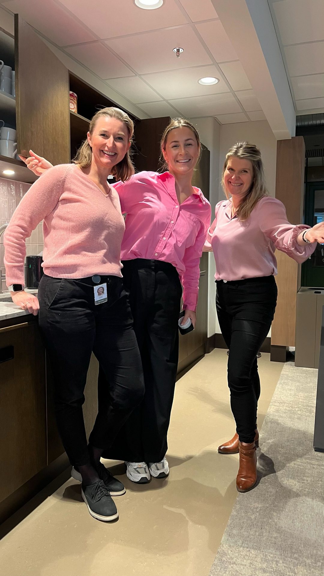 Three smiling women in pink tops and black pants pose in a modern kitchen setting.