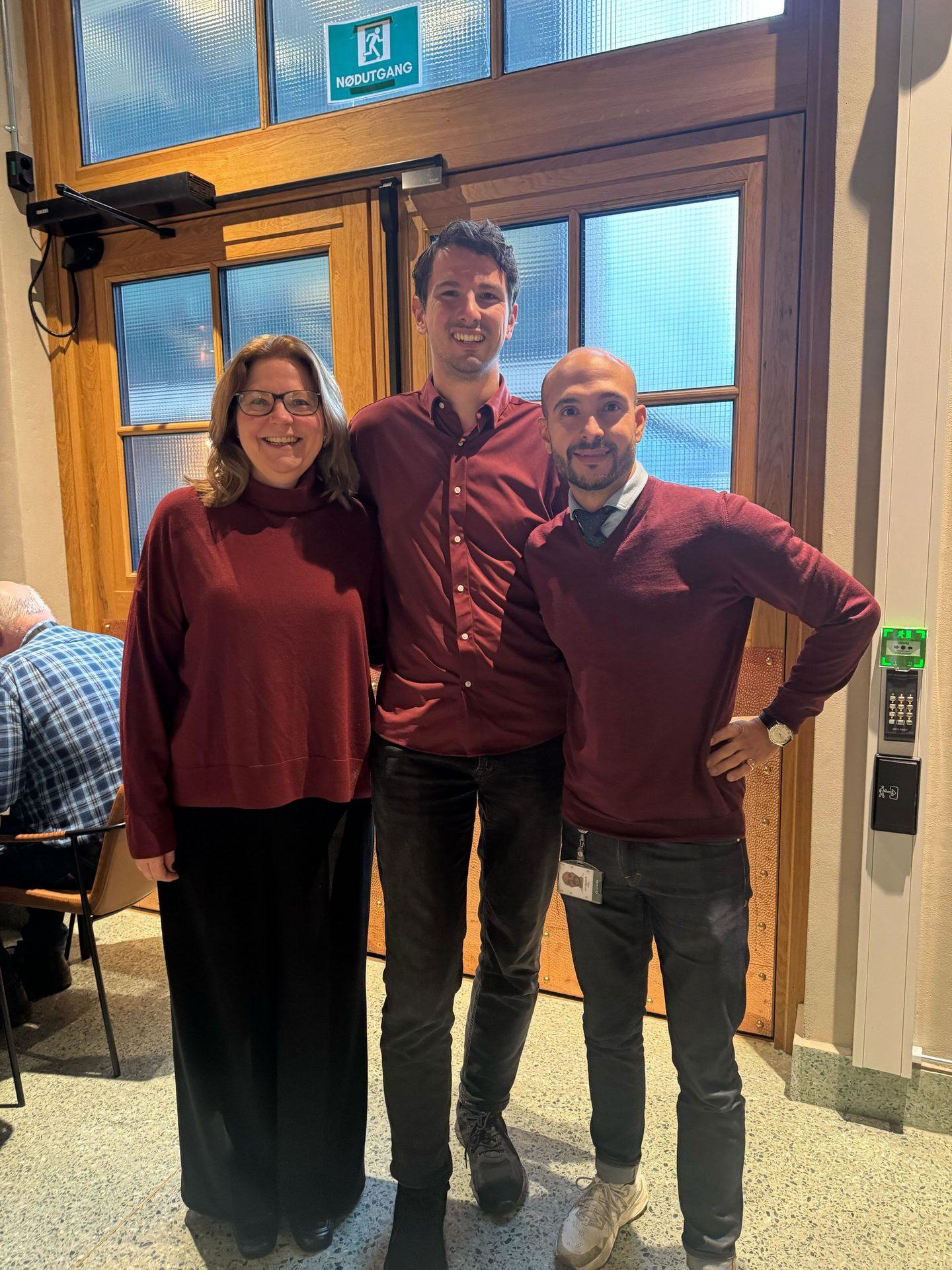 Three smiling people in maroon tops stand in front of a wooden door with an exit sign.