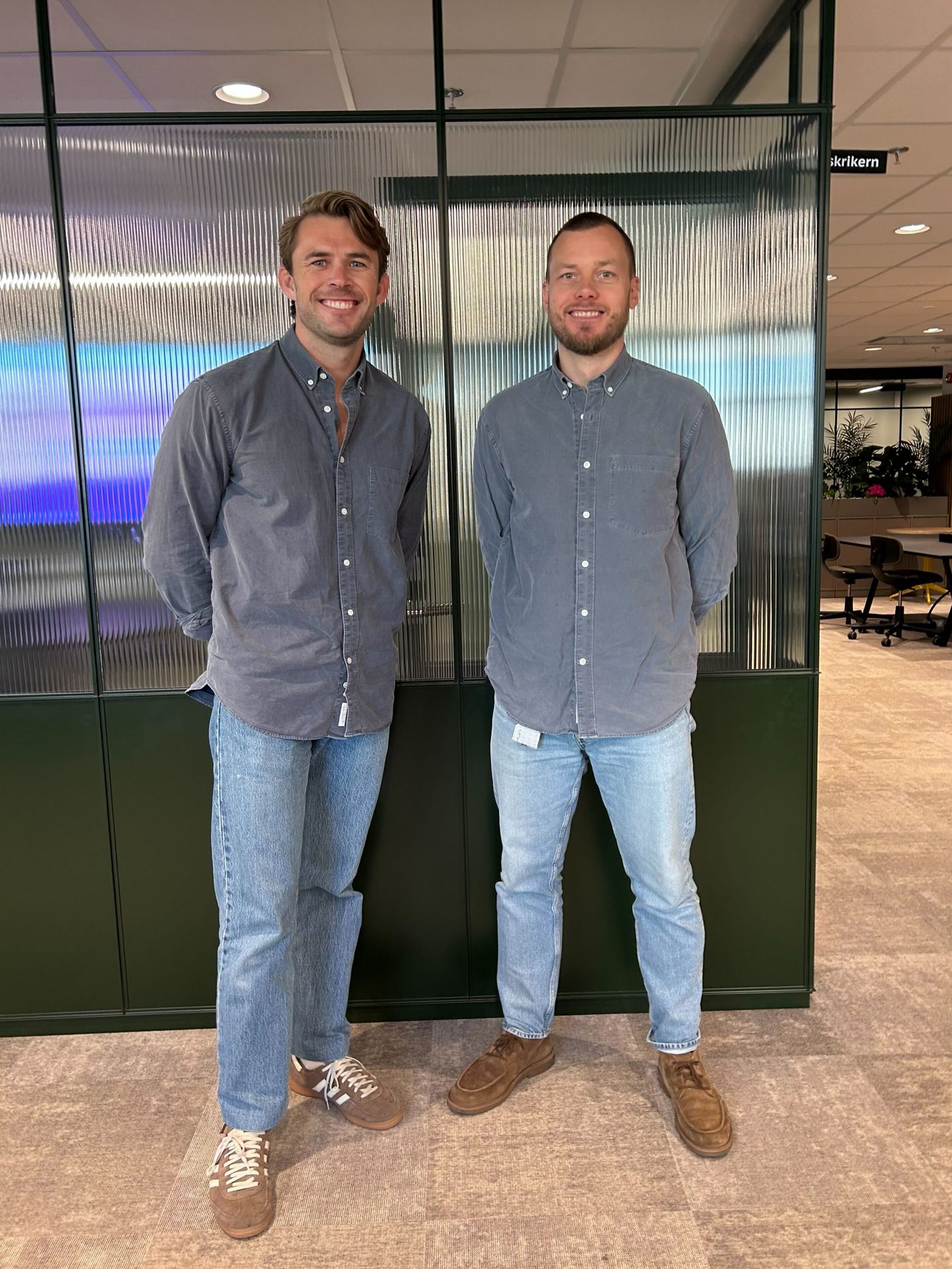 Two smiling men in gray shirts and blue jeans stand in an office with a fluted glass partition.