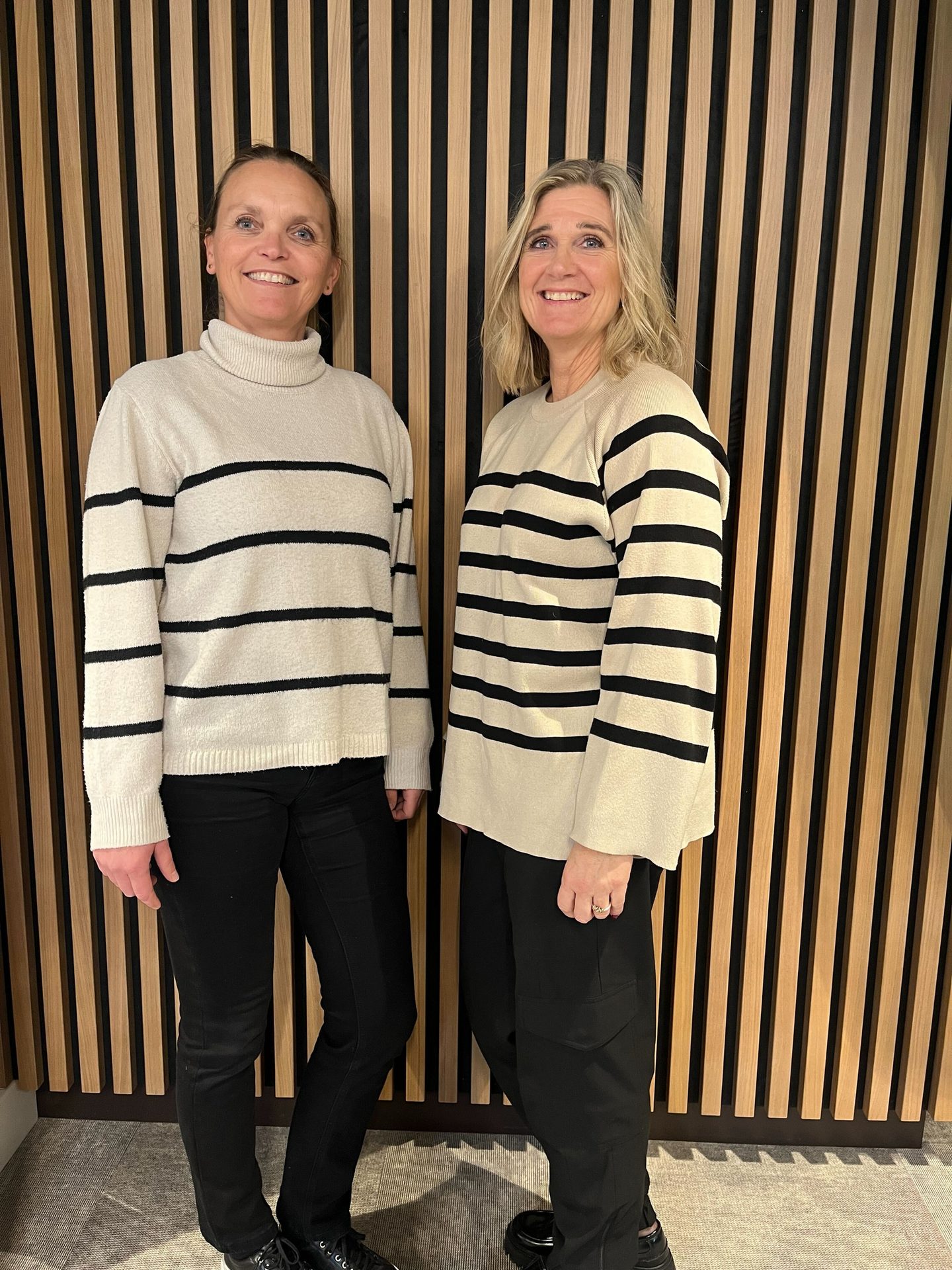 Two women in striped sweaters and dark pants stand against a slatted wooden wall, smiling at the camera.