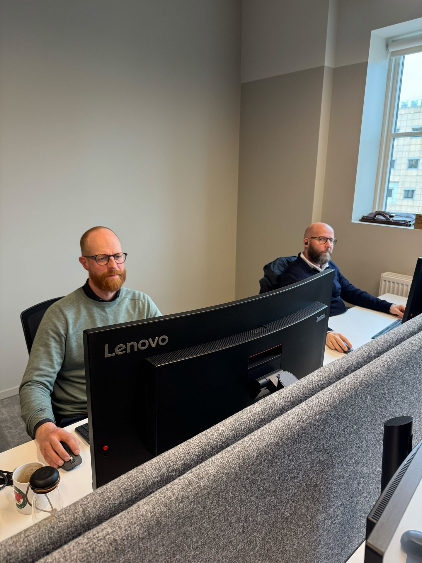 Two men working at computers in a modern office with desk dividers.