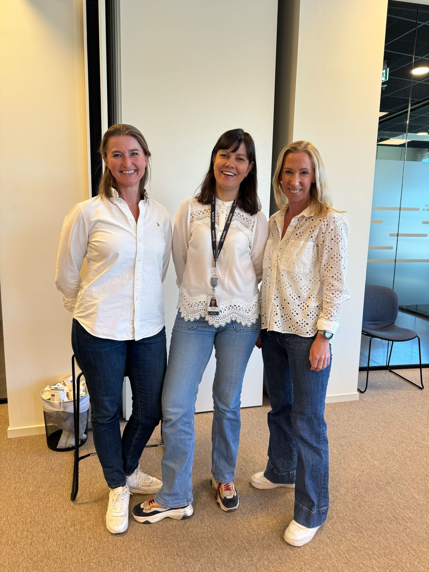Three smiling women in white tops and jeans stand together in an indoor setting.