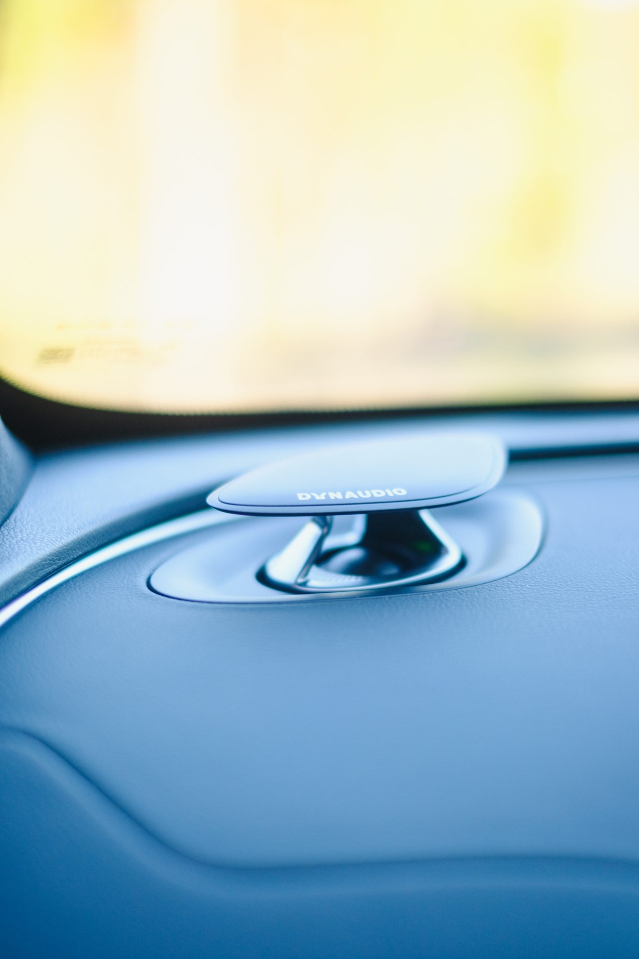 DYNAUDIO speaker on a car dashboard, with a blurred bright background.