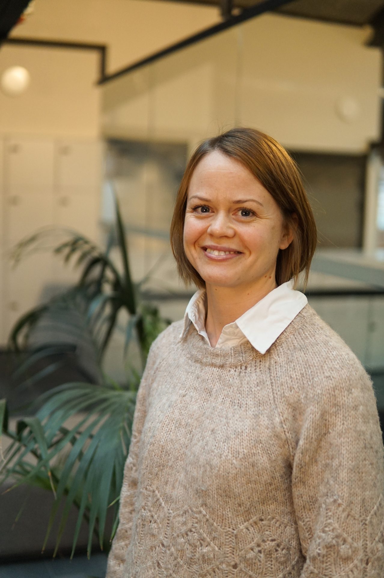A smiling woman with short brown hair, wearing a beige sweater and white shirt, in an office.