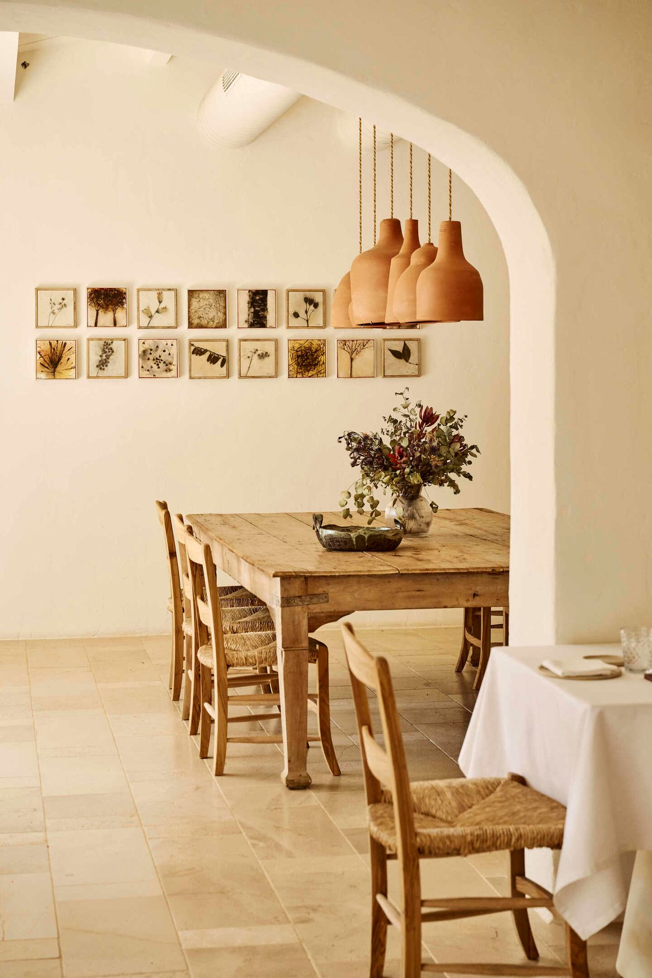 Rustic dining room with long wooden table, woven chairs, botanical prints, and terracotta pendants.