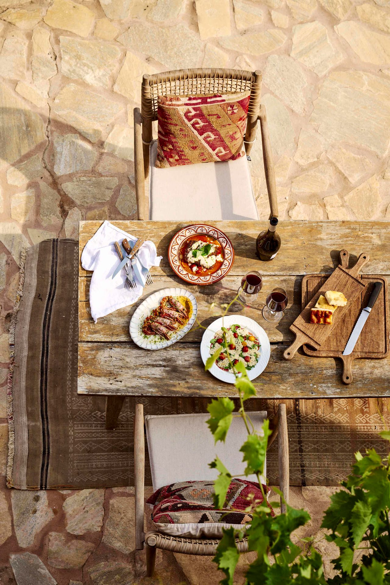 Rustic outdoor dining table with food, wine, and chairs on a stone patio.