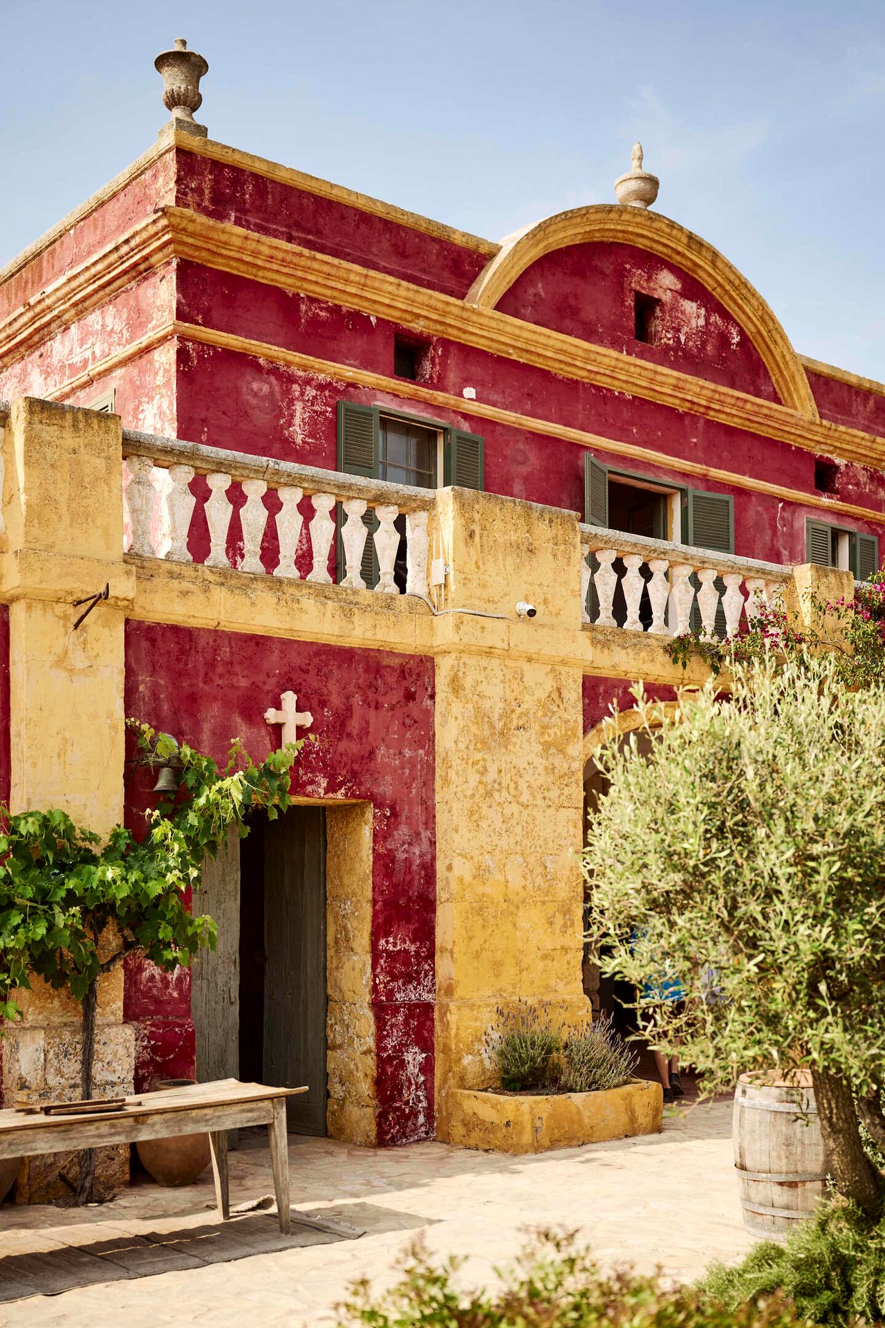 Rustic red and yellow building with a balcony, white cross, and olive tree.