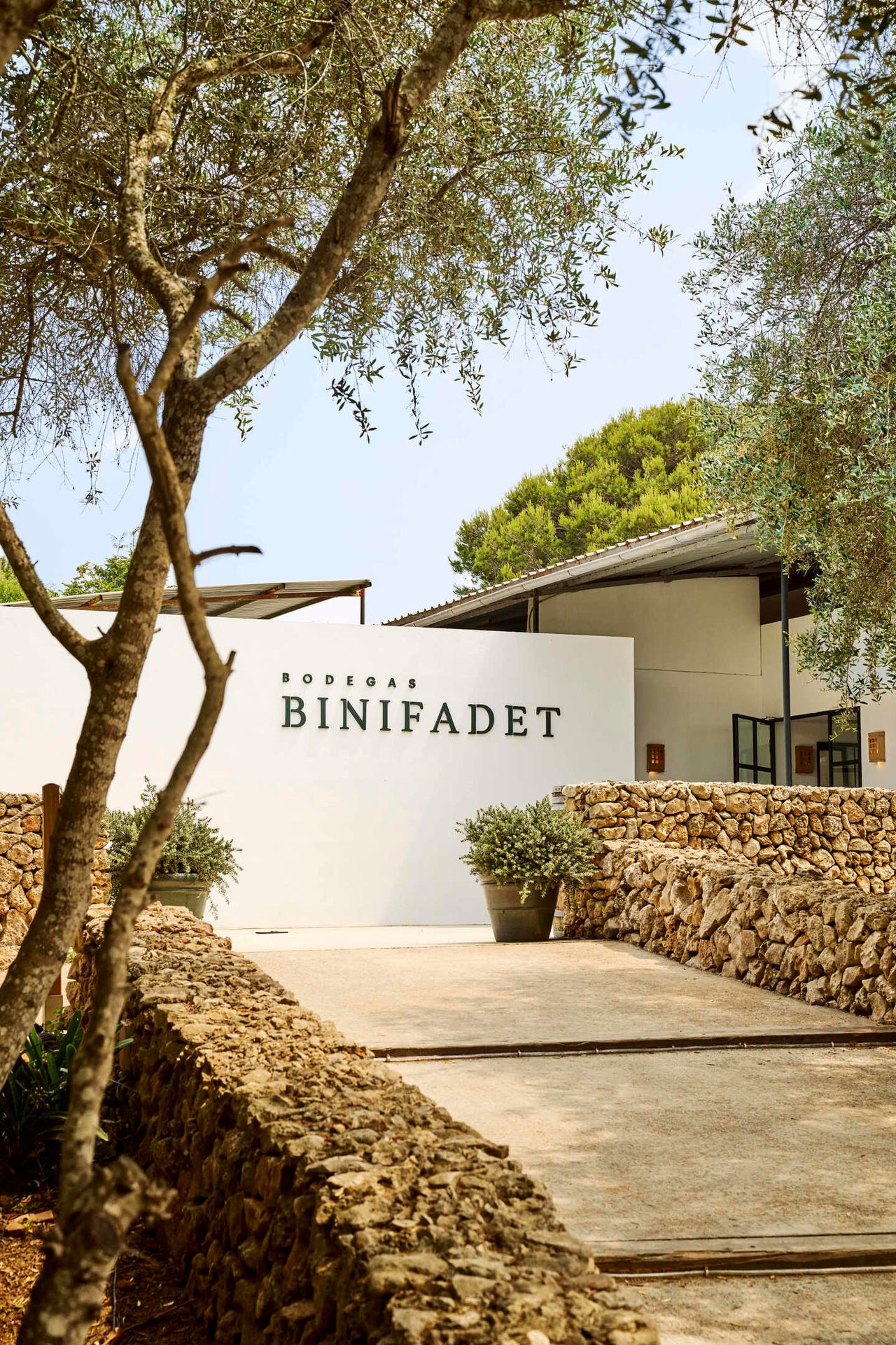 Entrance to Bodegas Binifadet winery, white building with sign, olive trees, and stone walls.
