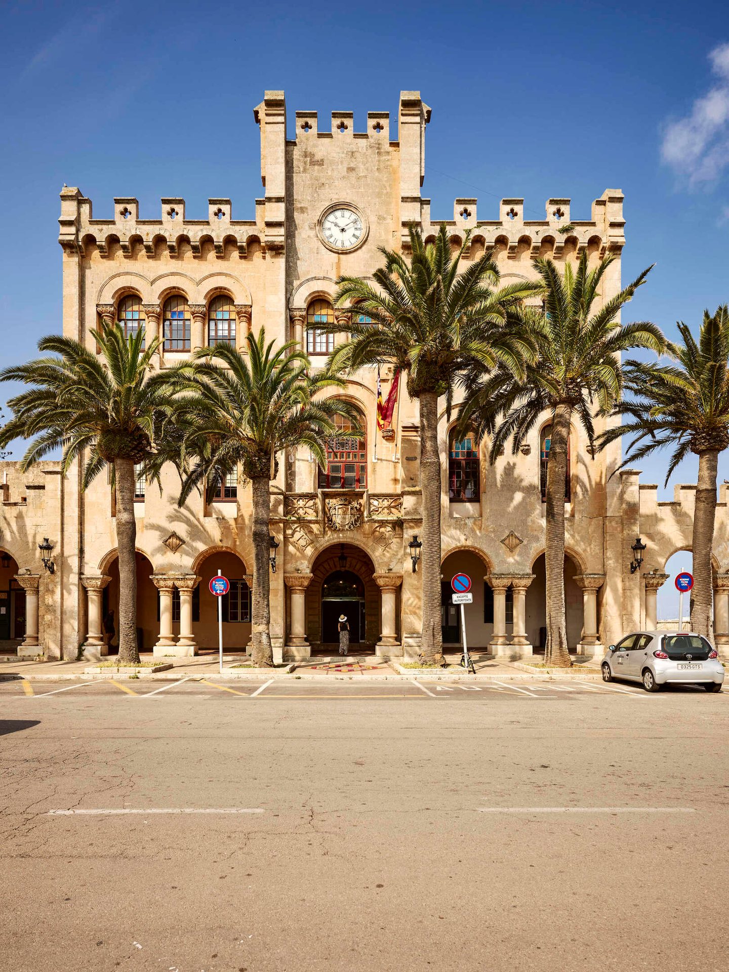 Historic stone building with clock tower, battlements, and palm trees on a street.