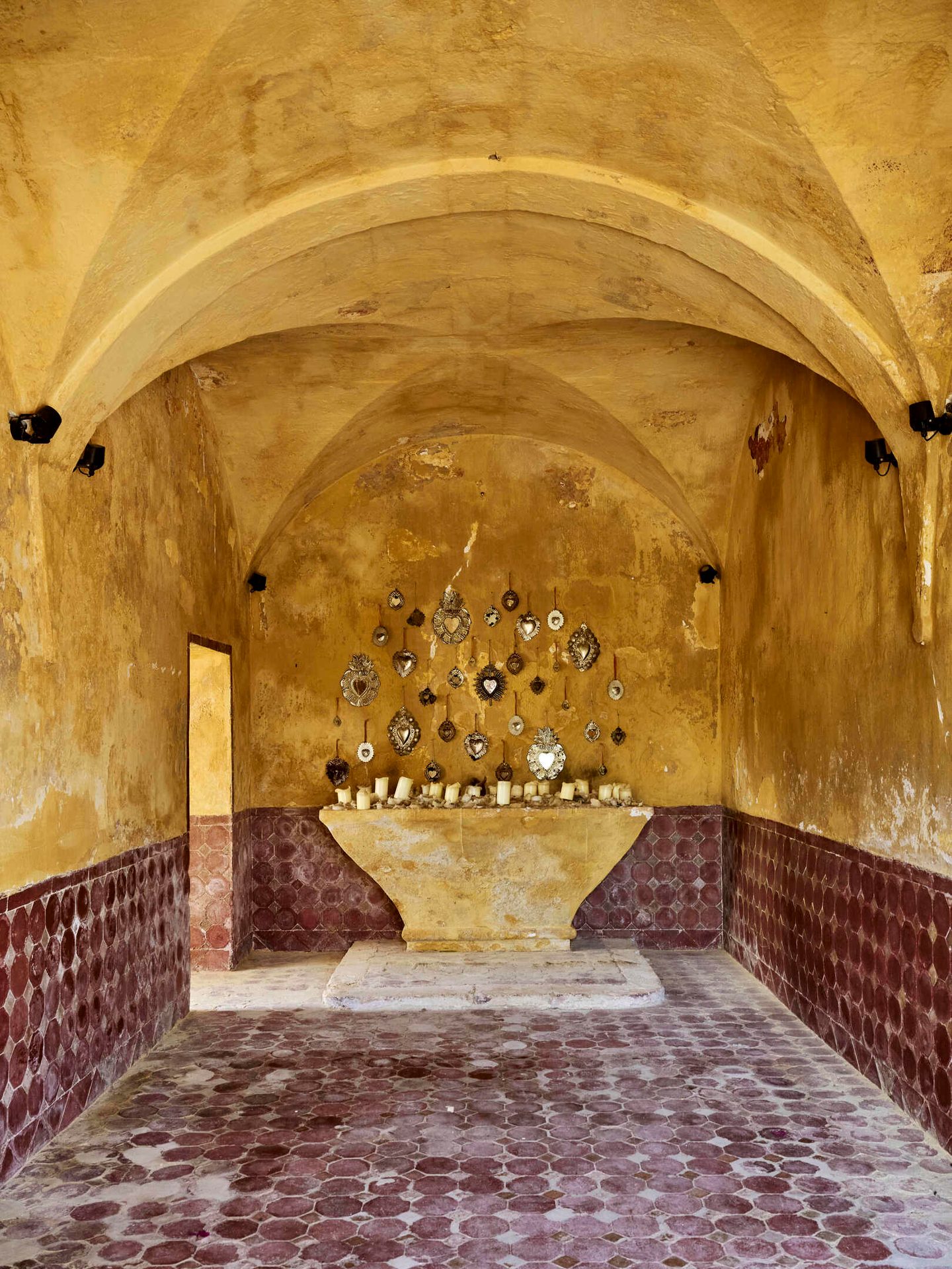 Arched yellow-ocher room, red hexagonal floor/wall tiles, stone altar with candles and silver hearts.