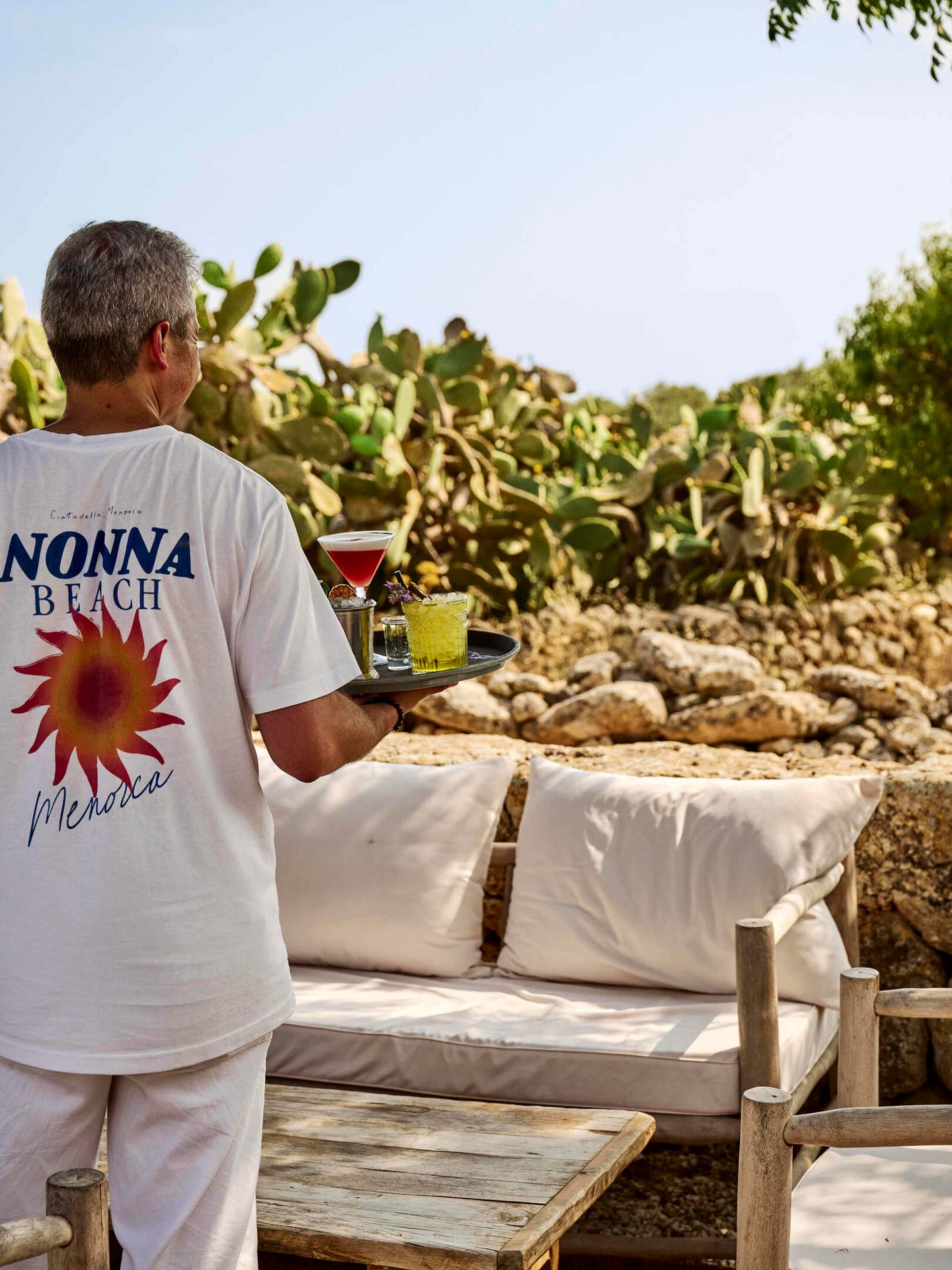 Waiter in "Nonna Beach Menorca" shirt carrying drinks on a tray outdoors with cacti.