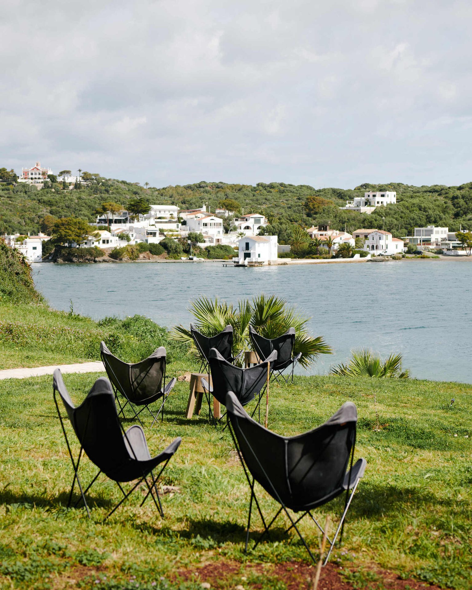 Butterfly chairs on grass overlooking a bay, with white houses on a distant green hillside.