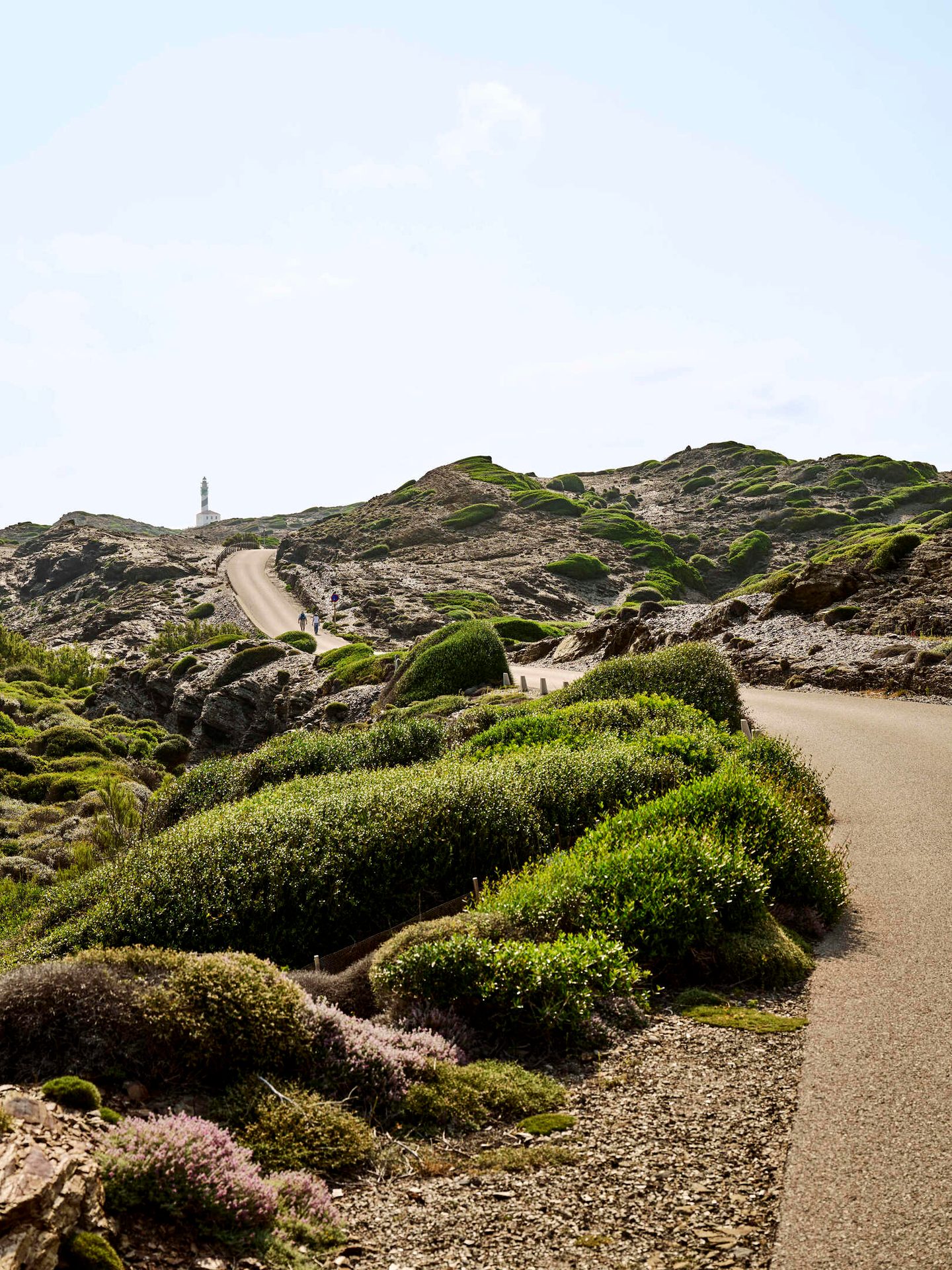 Winding road through rocky, green hills leads to a distant lighthouse, with people walking.