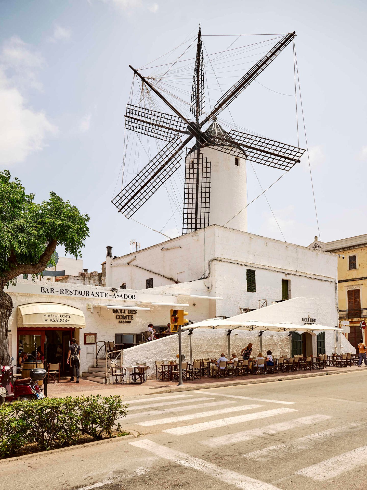 A large white windmill with grid sails stands above a white restaurant with outdoor diners on a sunny street.