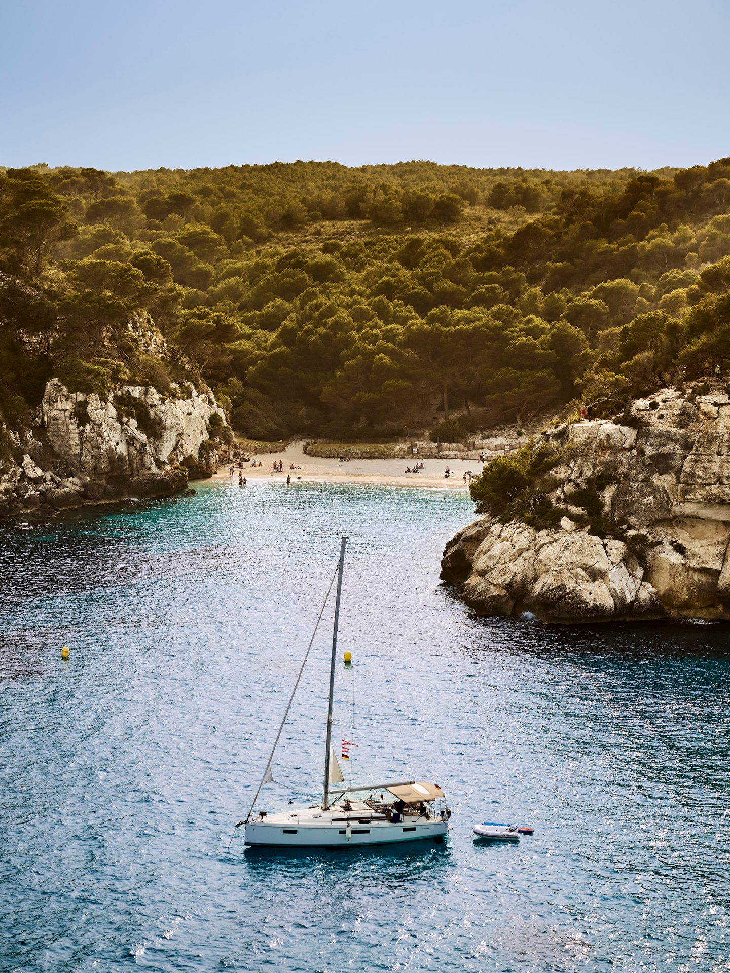 A sailboat anchored in a clear blue cove with a sandy beach, people, and tree-covered cliffs.