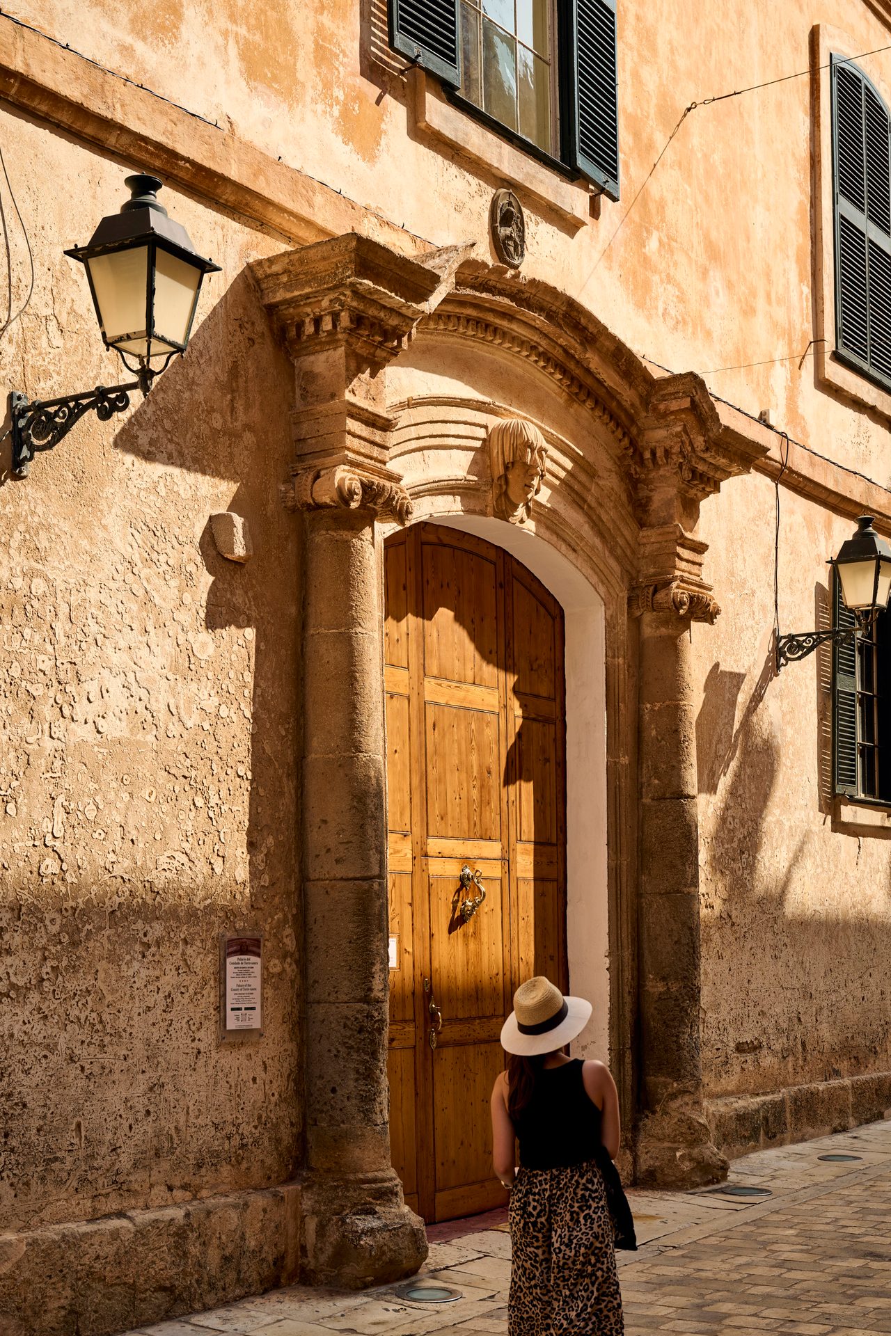Person in hat stands before ornate door of ancient building with strong shadows.