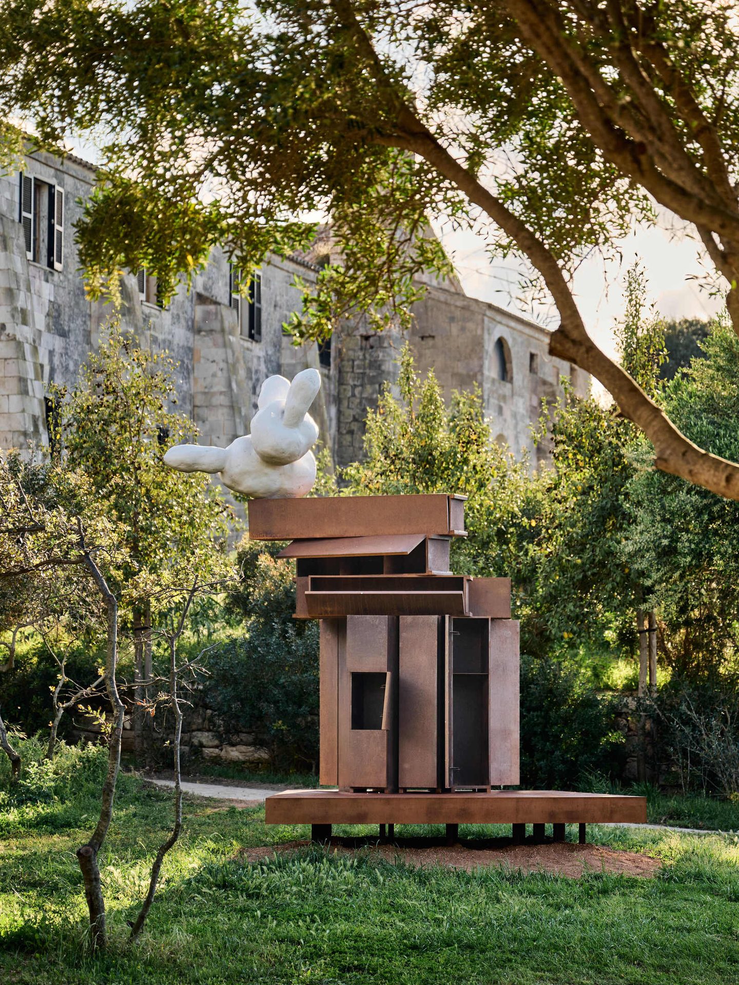White rabbit sculpture on rusty metal art in a green garden with an old stone building.