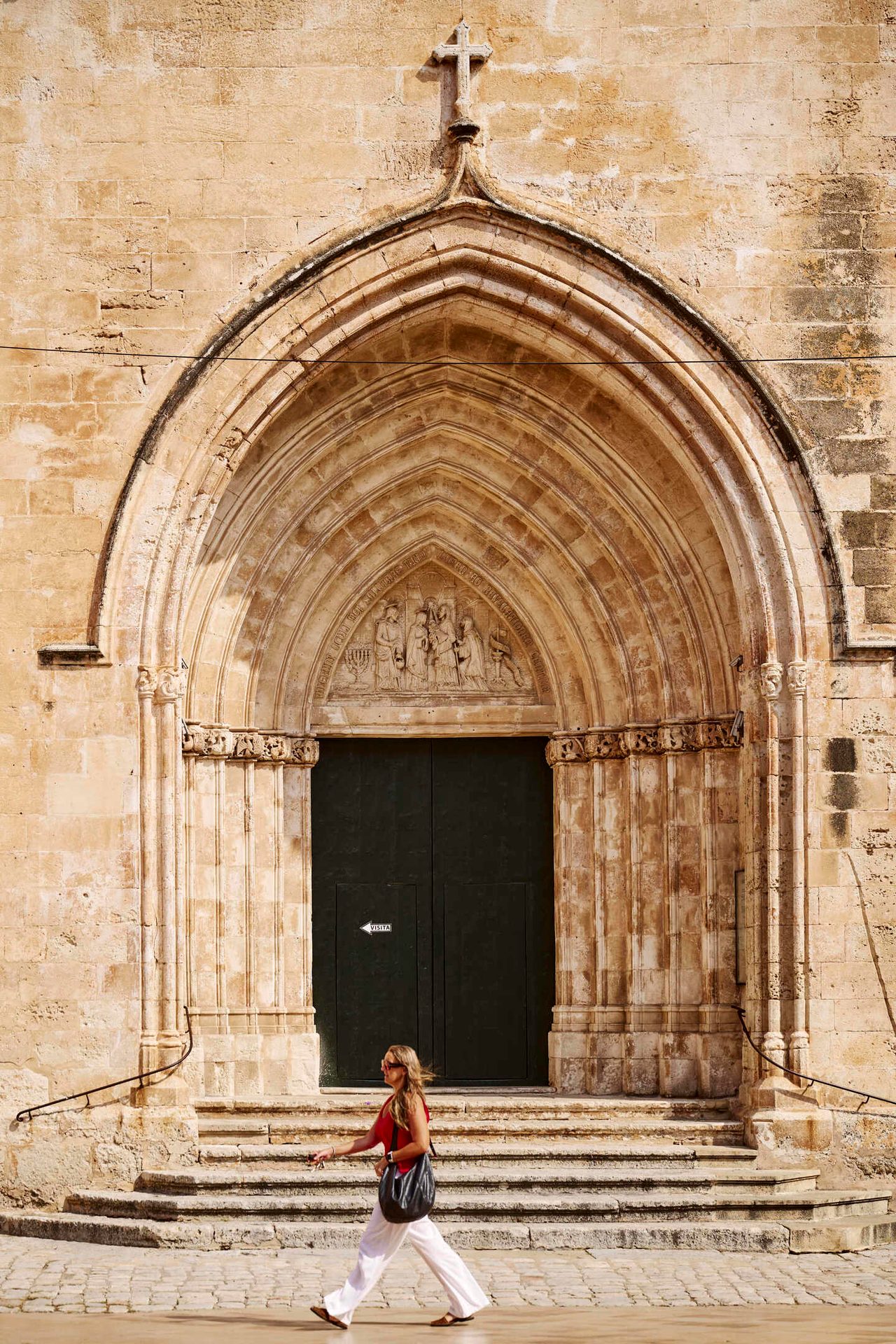 A woman walks past a stone church entrance with an archway and a cross.