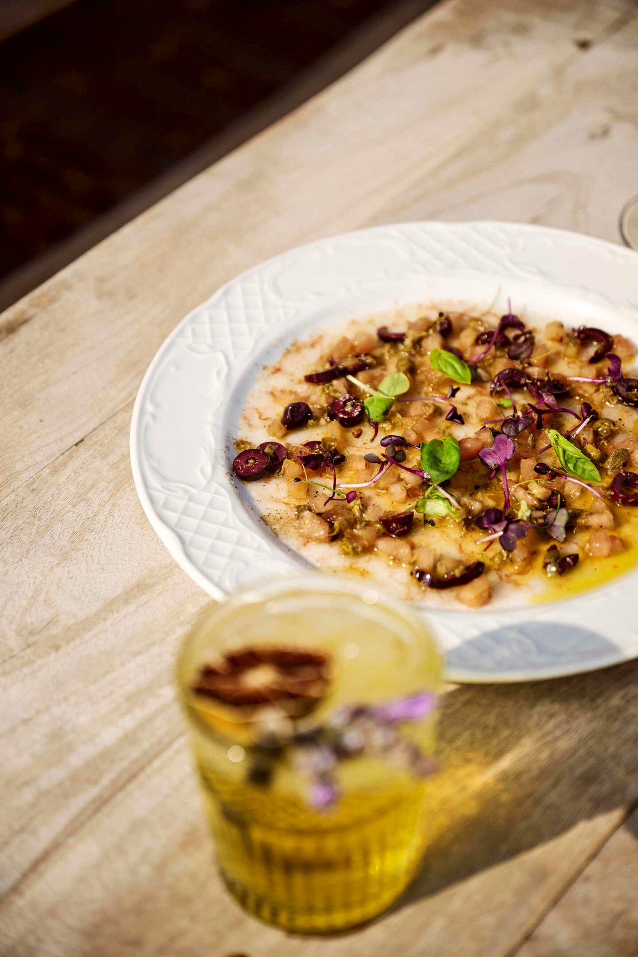Colorful fish carpaccio with olives & herbs on a white plate, next to a blurred drink.