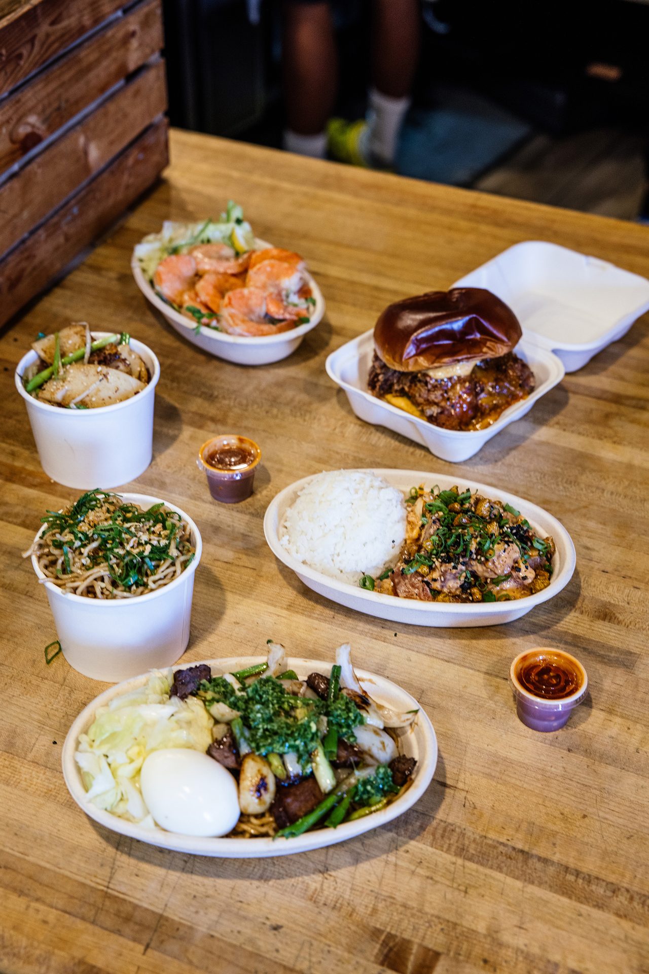 Various takeout food dishes including burger, shrimp, noodles, and rice on a table.