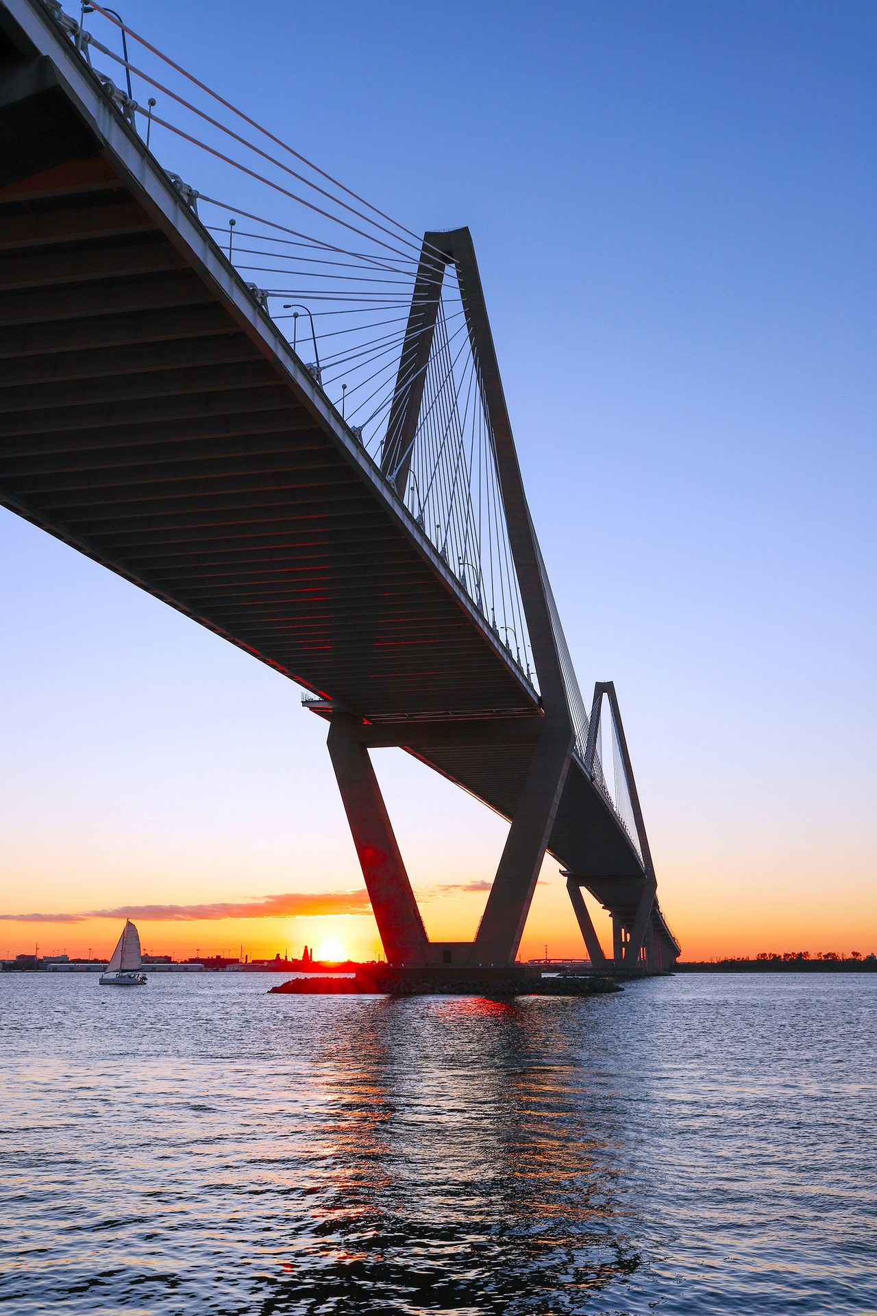 Sunset over a large bridge with a sailboat on the water.