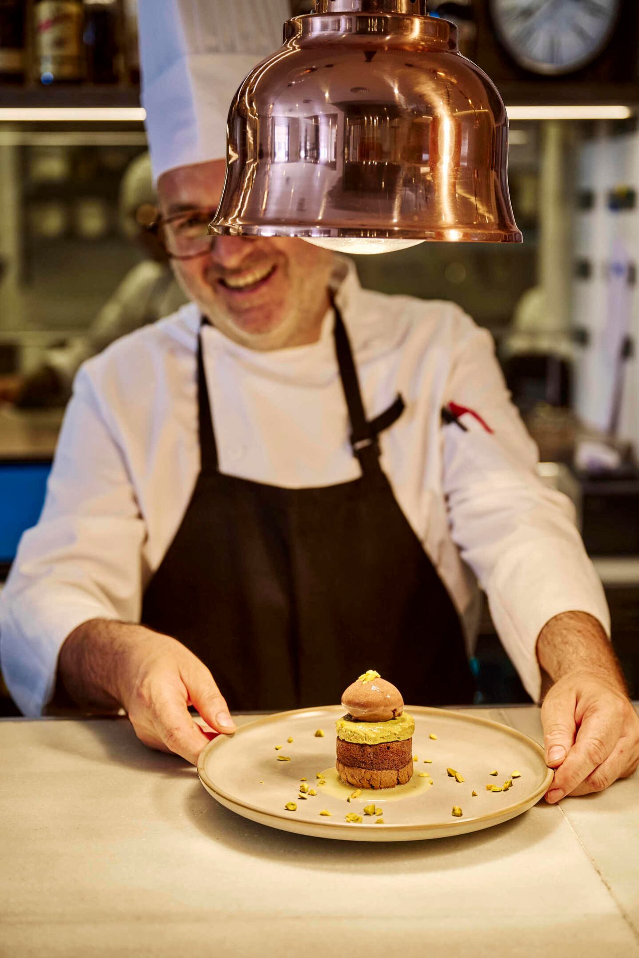 Smiling chef presenting a plated dessert under a copper lamp.