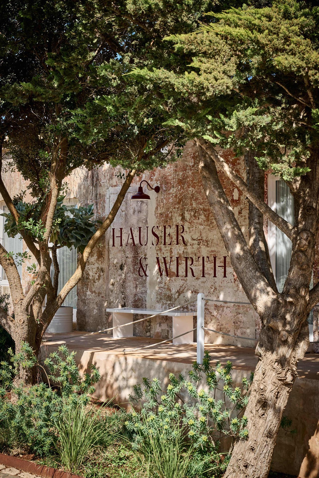 HAUSER & WIRTH sign on a rustic wall, framed by trees and plants.