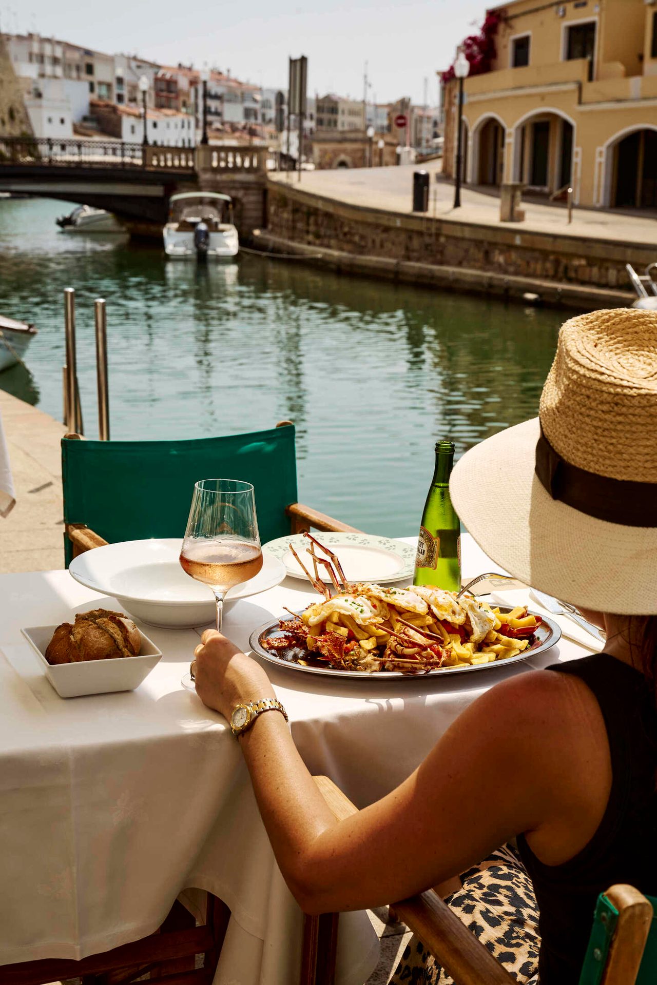 Woman in straw hat dining by canal with seafood, fries, and rosé wine.
