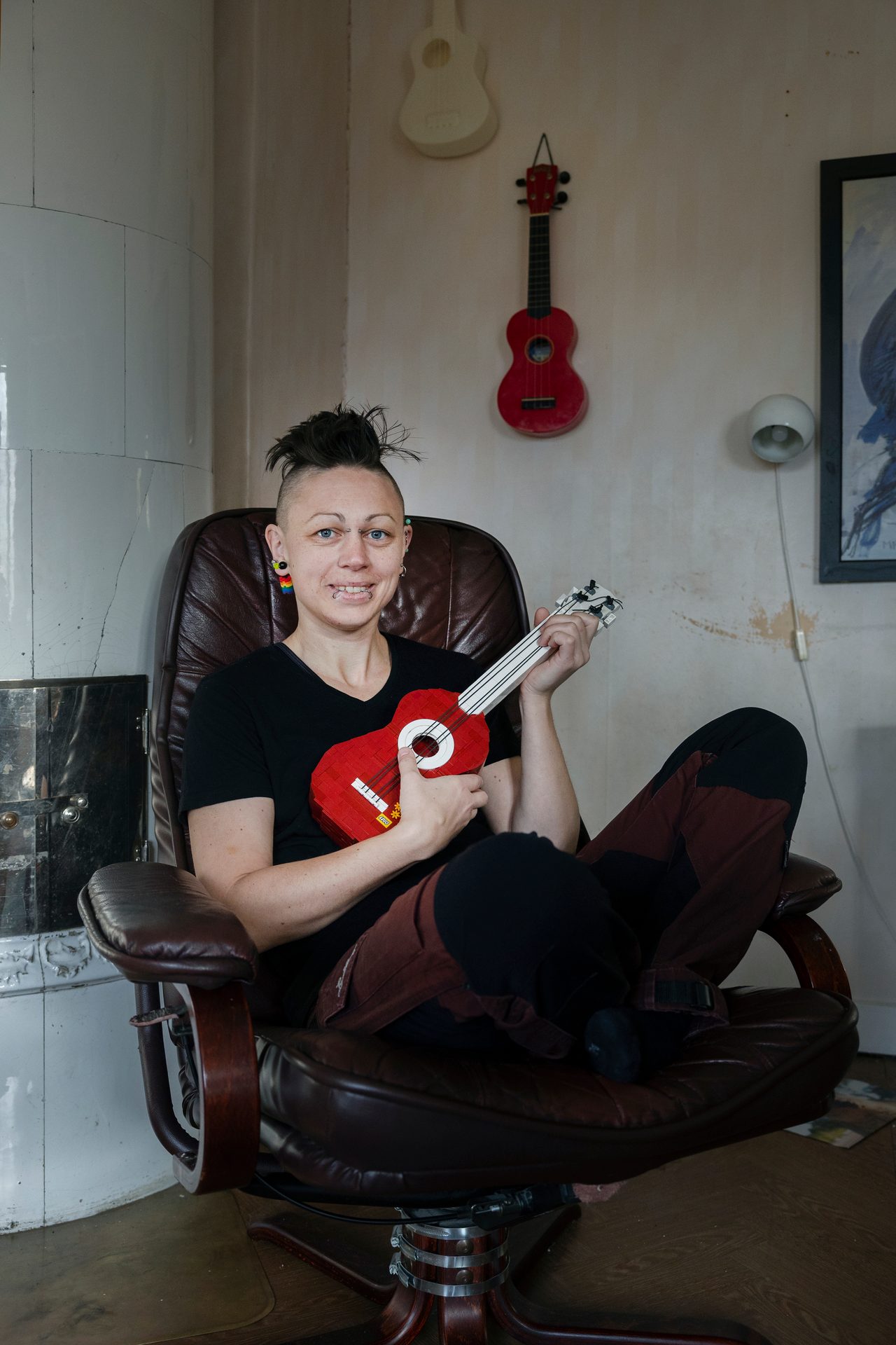A person with spiky hair and piercings holds a red LEGO ukulele while sitting in a chair.