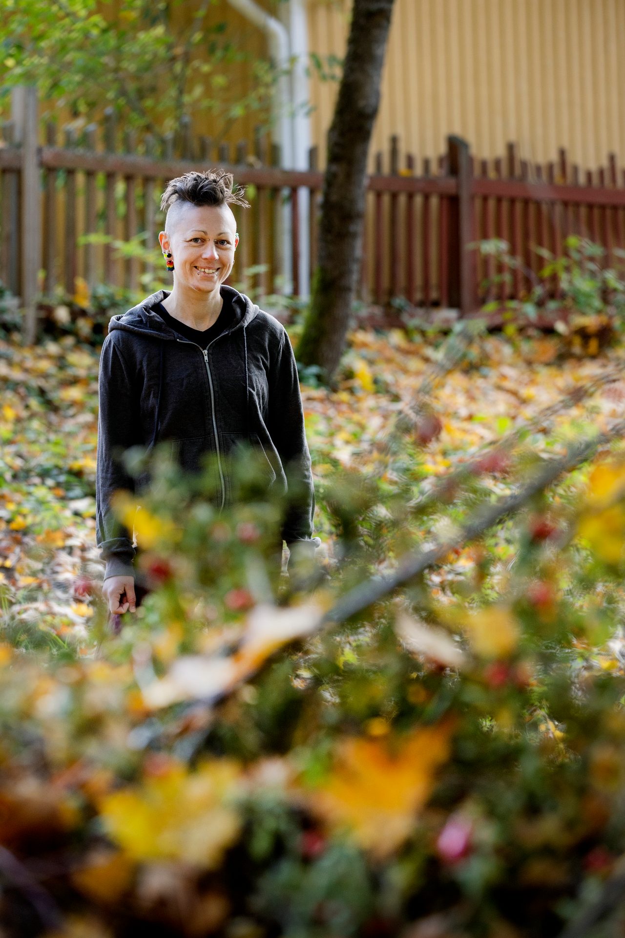 A smiling person with spiky hair in a dark hoodie stands amidst autumn leaves outdoors.