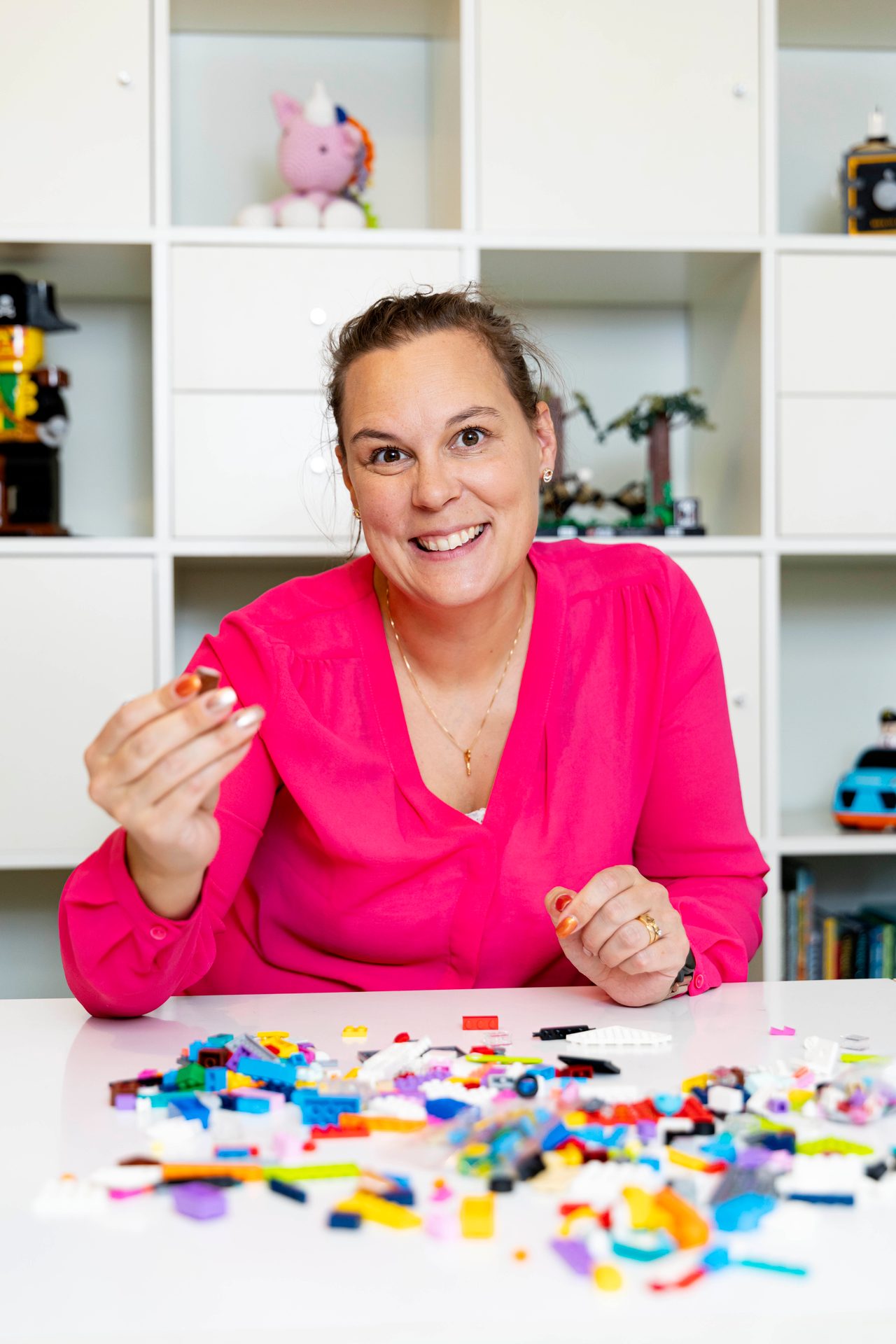 Smiling woman in pink holds a Lego piece, surrounded by colorful bricks on a white table.