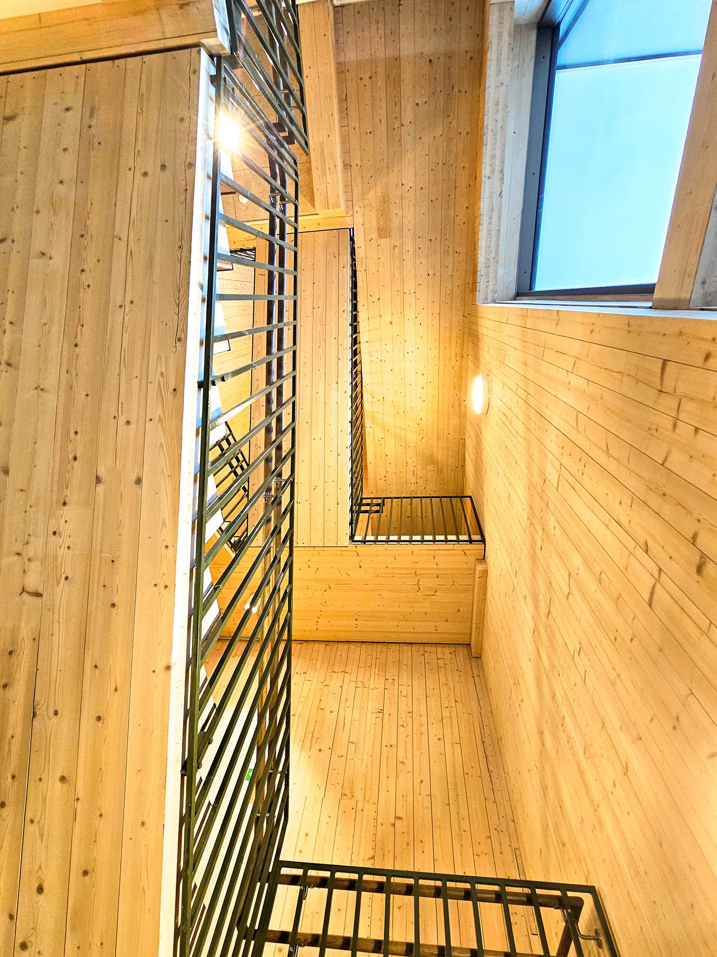 A multi-level wooden interior with metal railings, a large window, and wall-mounted light.