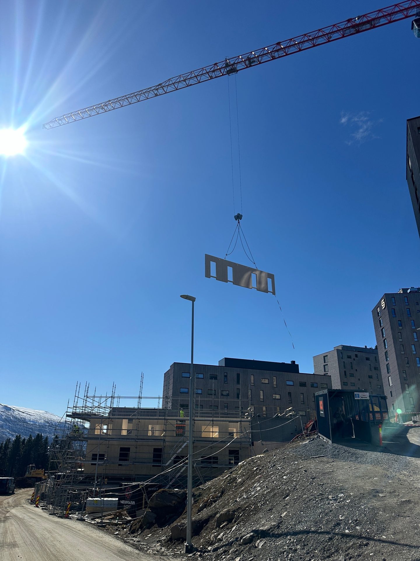 Crane lifts building panel on sunny construction site with new apartments and mountains.