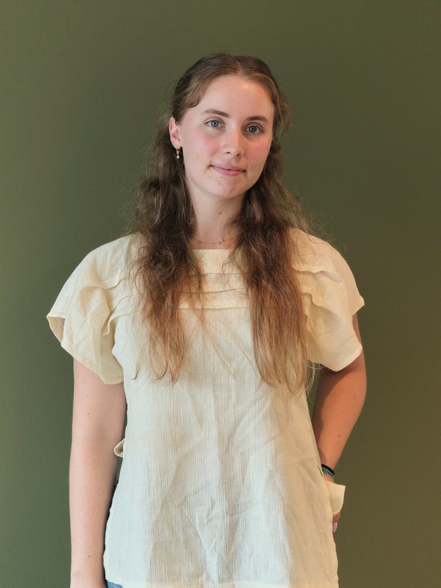 Young woman with long brown hair, blue eyes, wearing a yellow top against a green wall.
