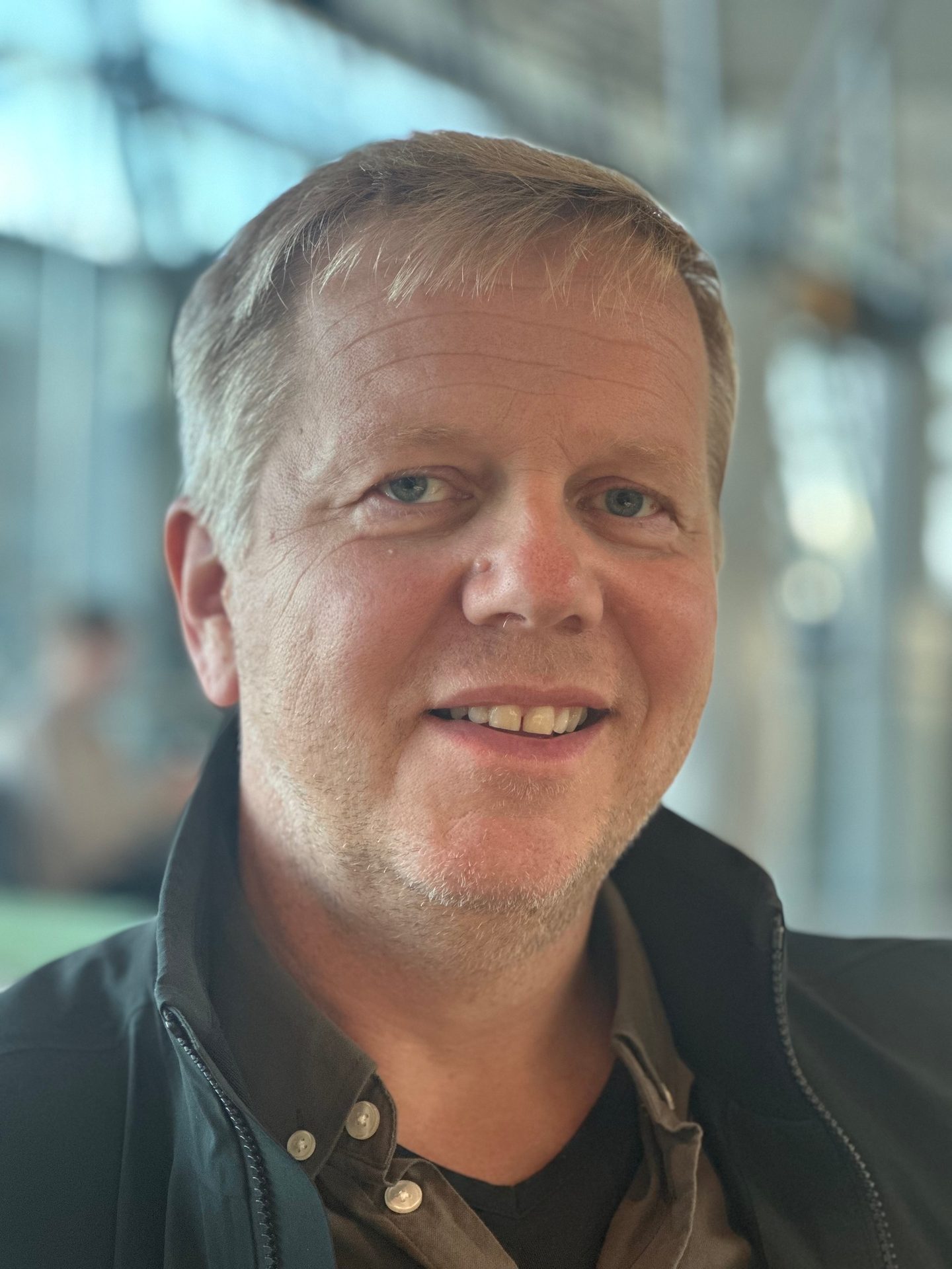 A smiling man with light hair and blue eyes wears a dark jacket, looking directly at the viewer.
