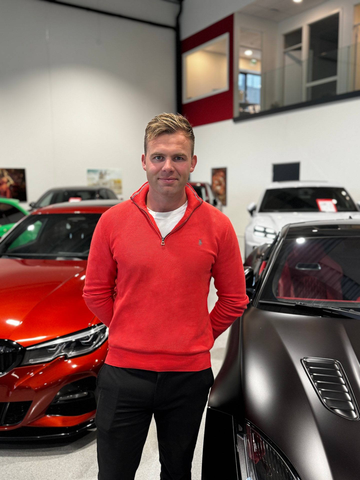 Man in a red sweater standing between a red and a black luxury car in a car dealership showroom.
