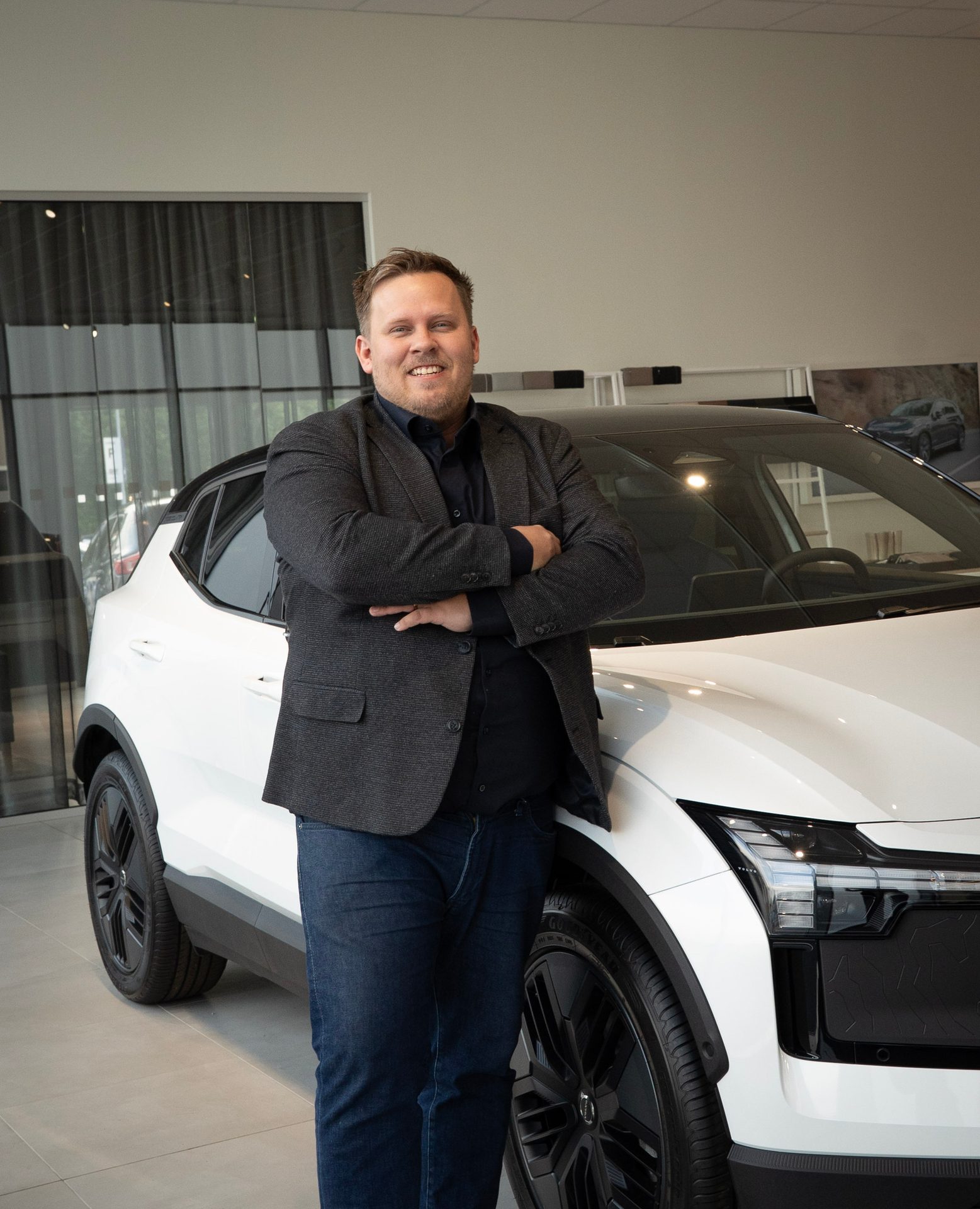 Smiling man in a suit jacket and jeans, arms crossed, leaning against a white electric car.