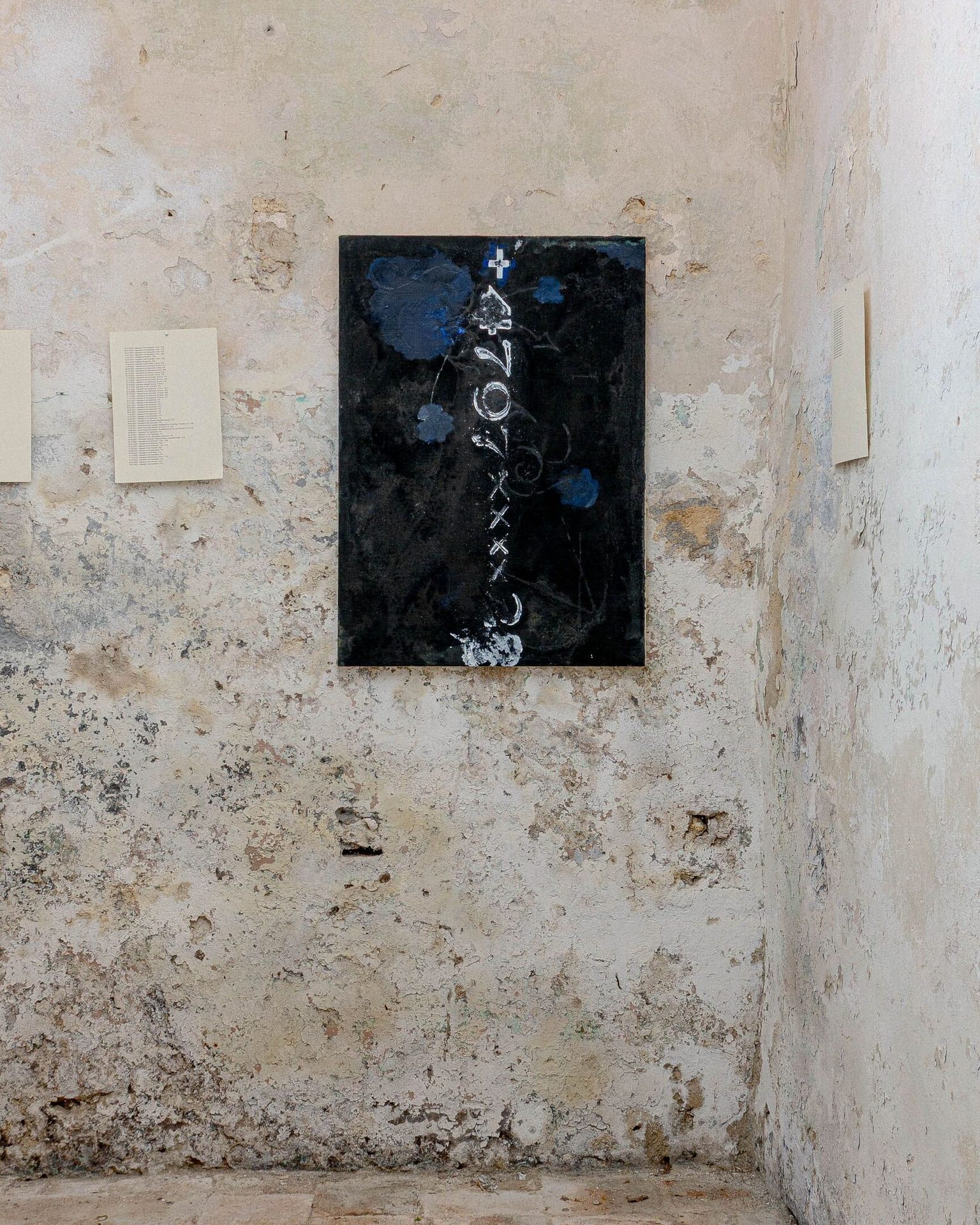 Vertical black painting with white symbols & blue flowers on a peeling, cracked wall.
