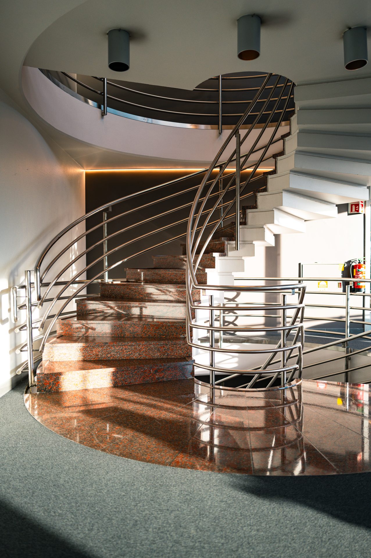 Modern spiral staircase with granite and white steps, silver railings, and a shiny reflective floor.