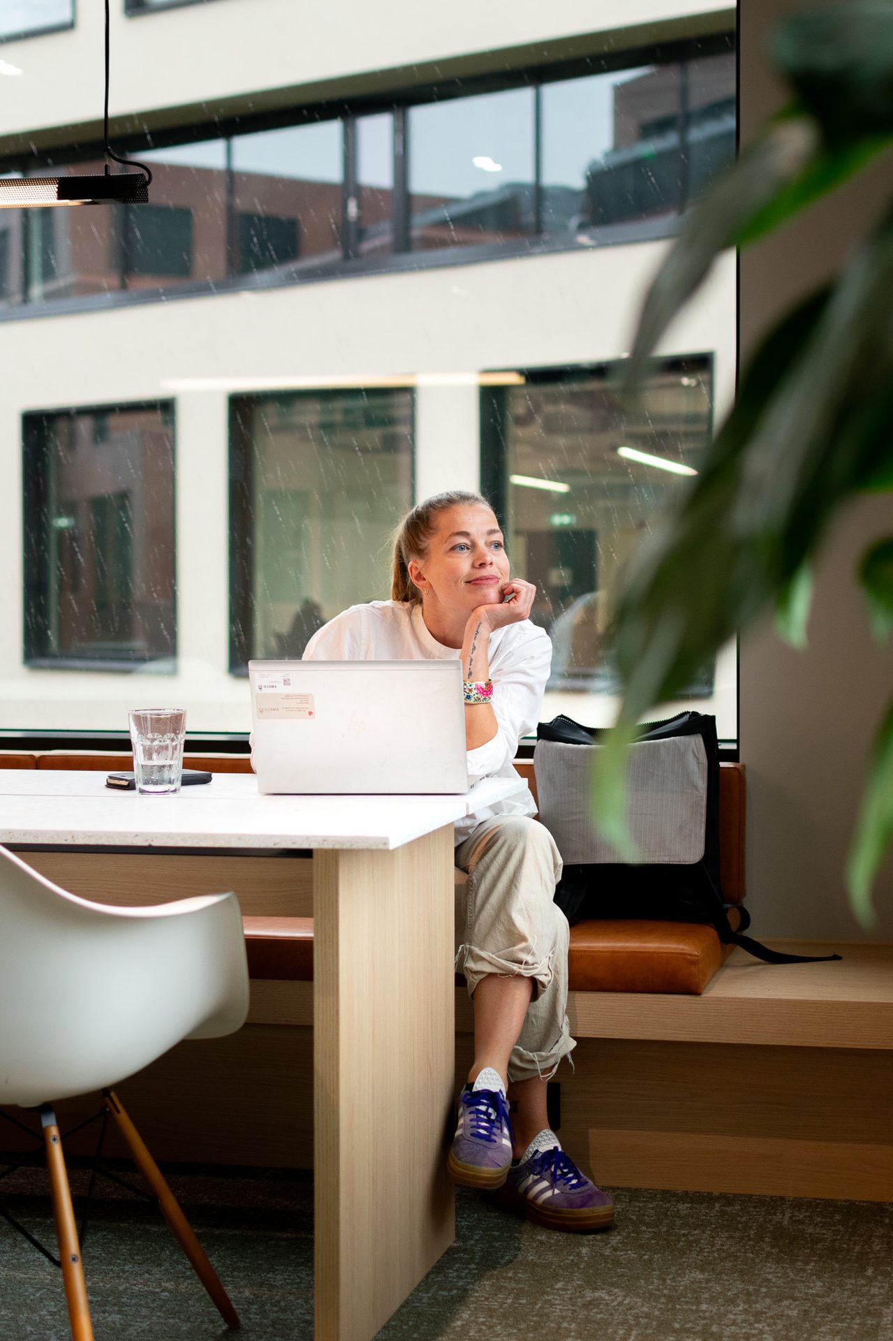 Woman working on a laptop, looking out a window in a modern workspace.