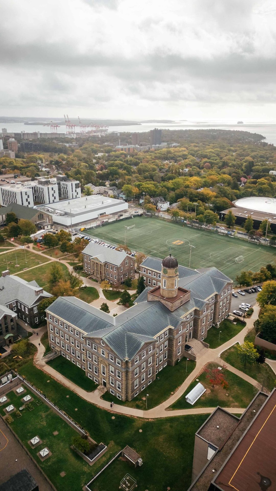 Aerial view of a university campus with stone buildings, clock tower, sports field, and harbor.