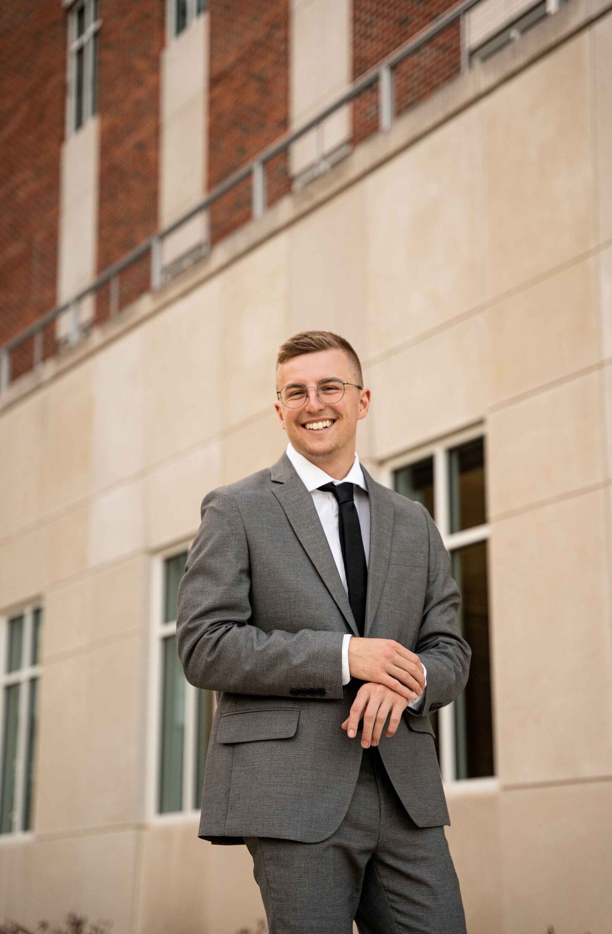 A young man in a gray suit and glasses smiles, standing confidently in front of a modern building.