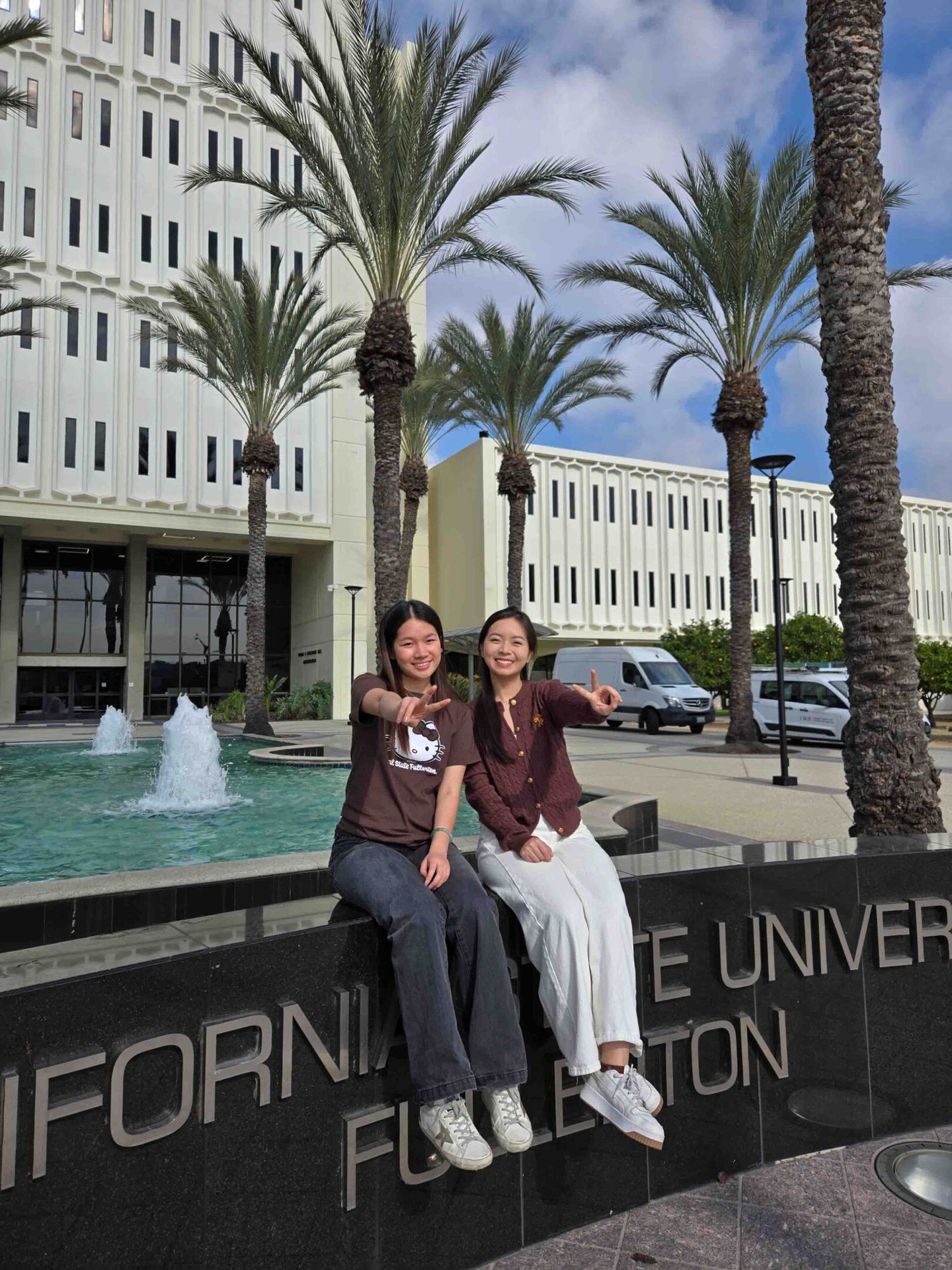 Two young women posing on a stone wall in front of a building with palm trees and a fountain.